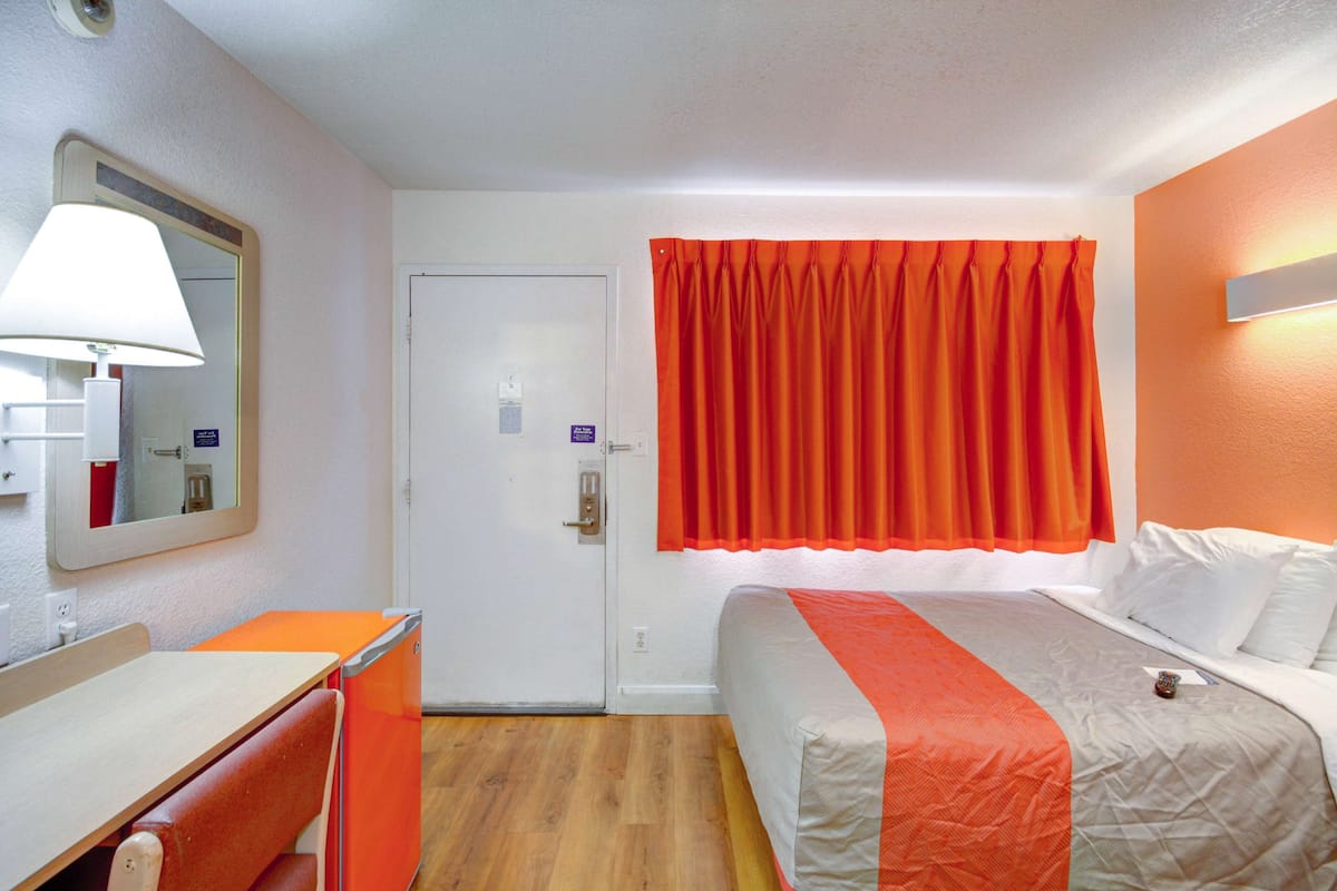 A hotel room with a bed, a desk, a chair, a mirror, and a door.