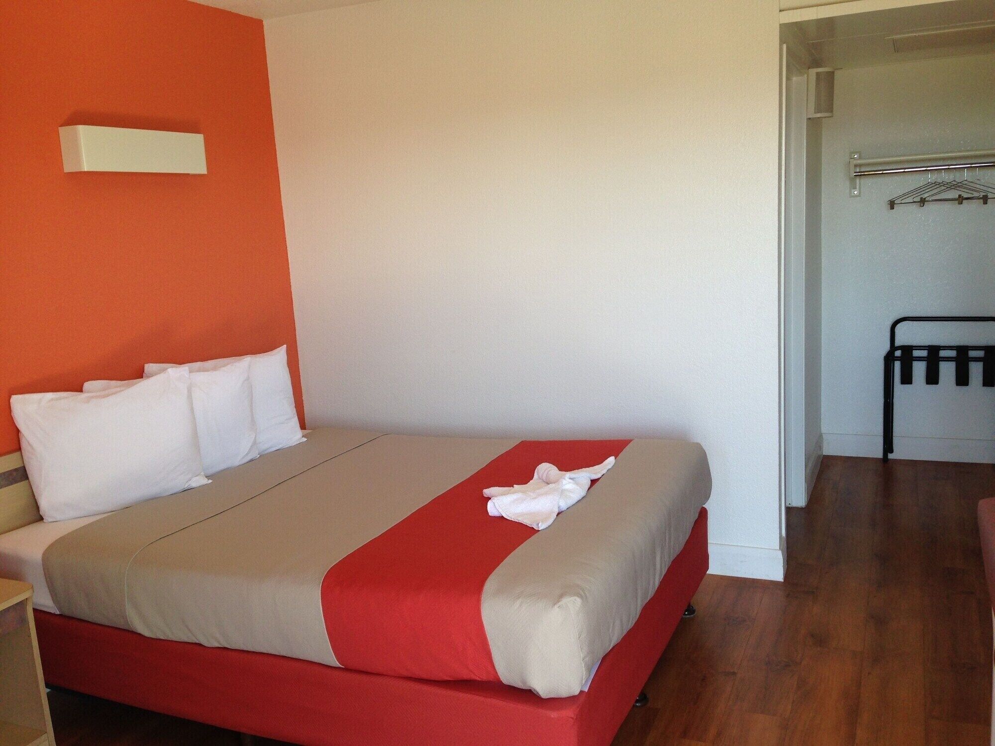 Una habitación de hotel con una cama, una colcha roja y beige, almohadas blancas y suelo de madera.