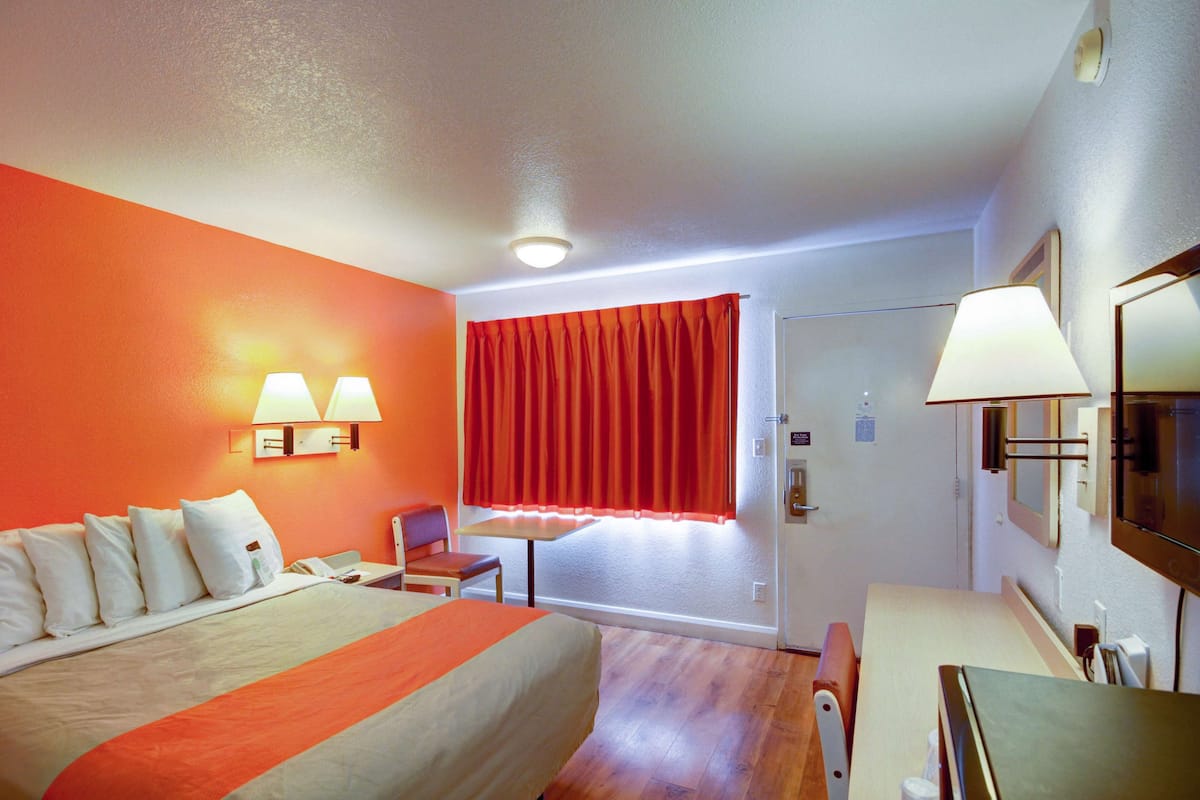 A hotel room with a bed, a desk, a chair, a window with red curtains, and wall-mounted lamps.