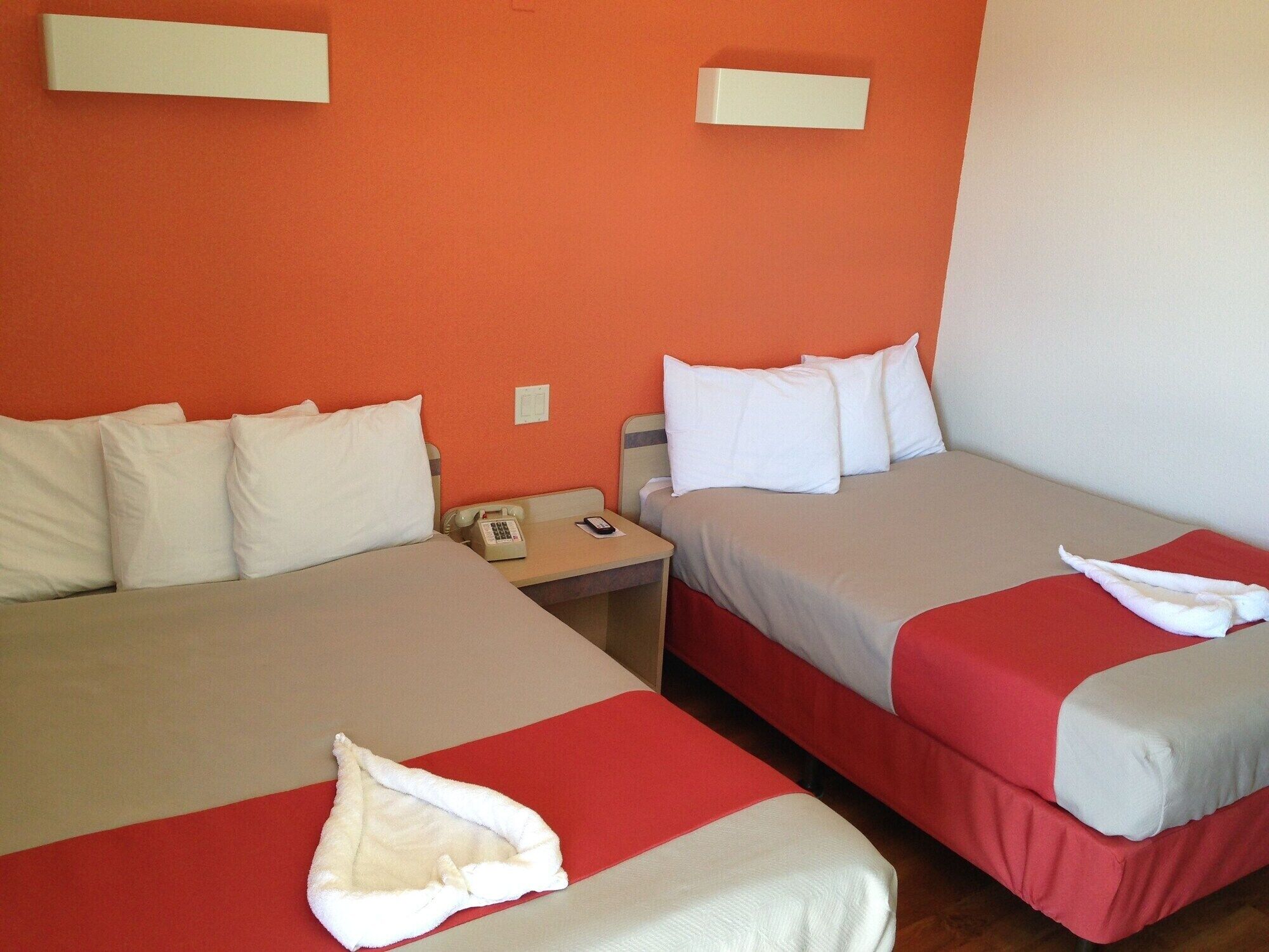 A hotel room with two beds, each with white bedding and red accents. There is a bedside table with a telephone and a lamp.