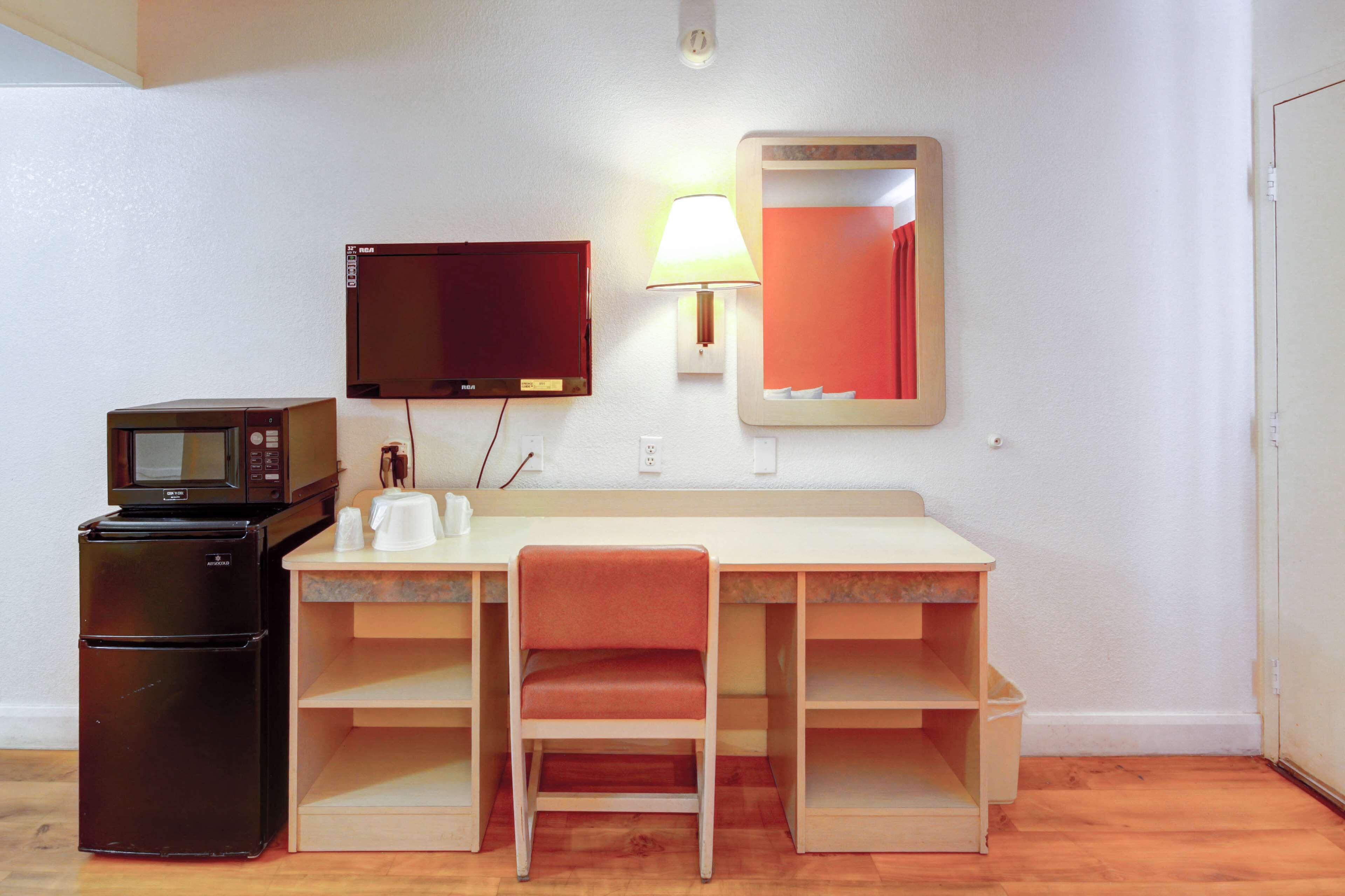 Habitación compacta con kitchenette, escritorio y silla, televisor de pantalla plana, lámpara y espejo.
