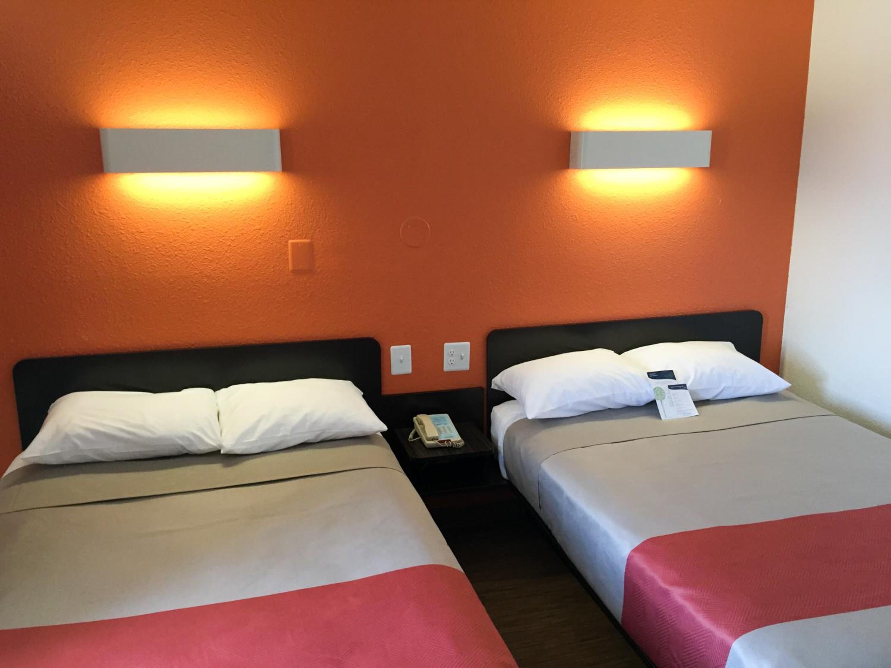 A hotel room with two beds, each with white pillows and a red blanket. There are wall-mounted lights above the beds and a phone on the nightstand.
