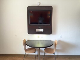 Una habitación con televisión de pantalla plana montada en la pared, una mesita pequeña y dos sillas.