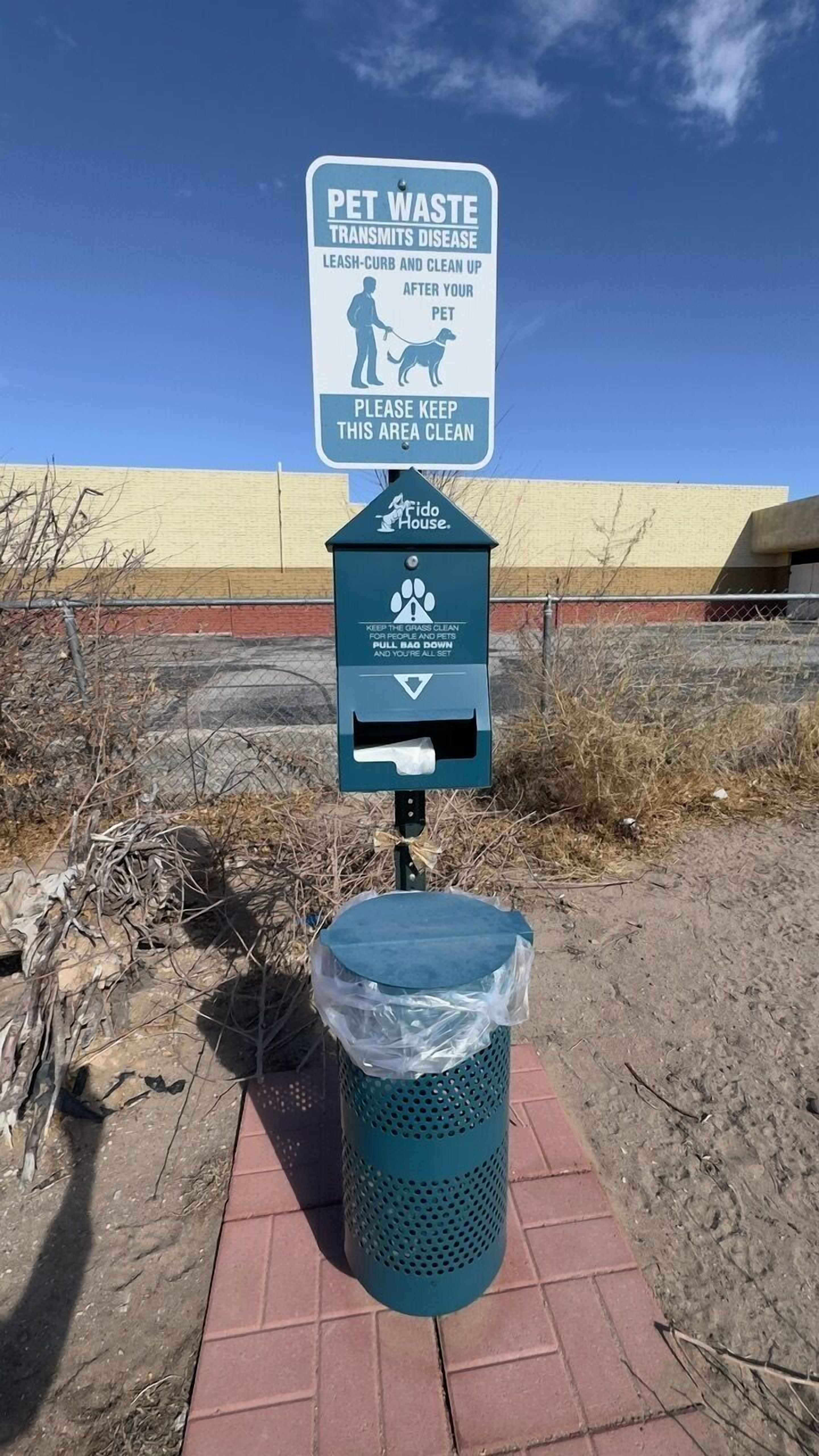 A pet waste station with a sign warning about disease transmission, a trash bin, and a fenced area.