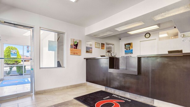 A reception area with a dark reception desk, a welcome mat, and a clock on the wall. - Motel 6 Grants, NM (Grants)