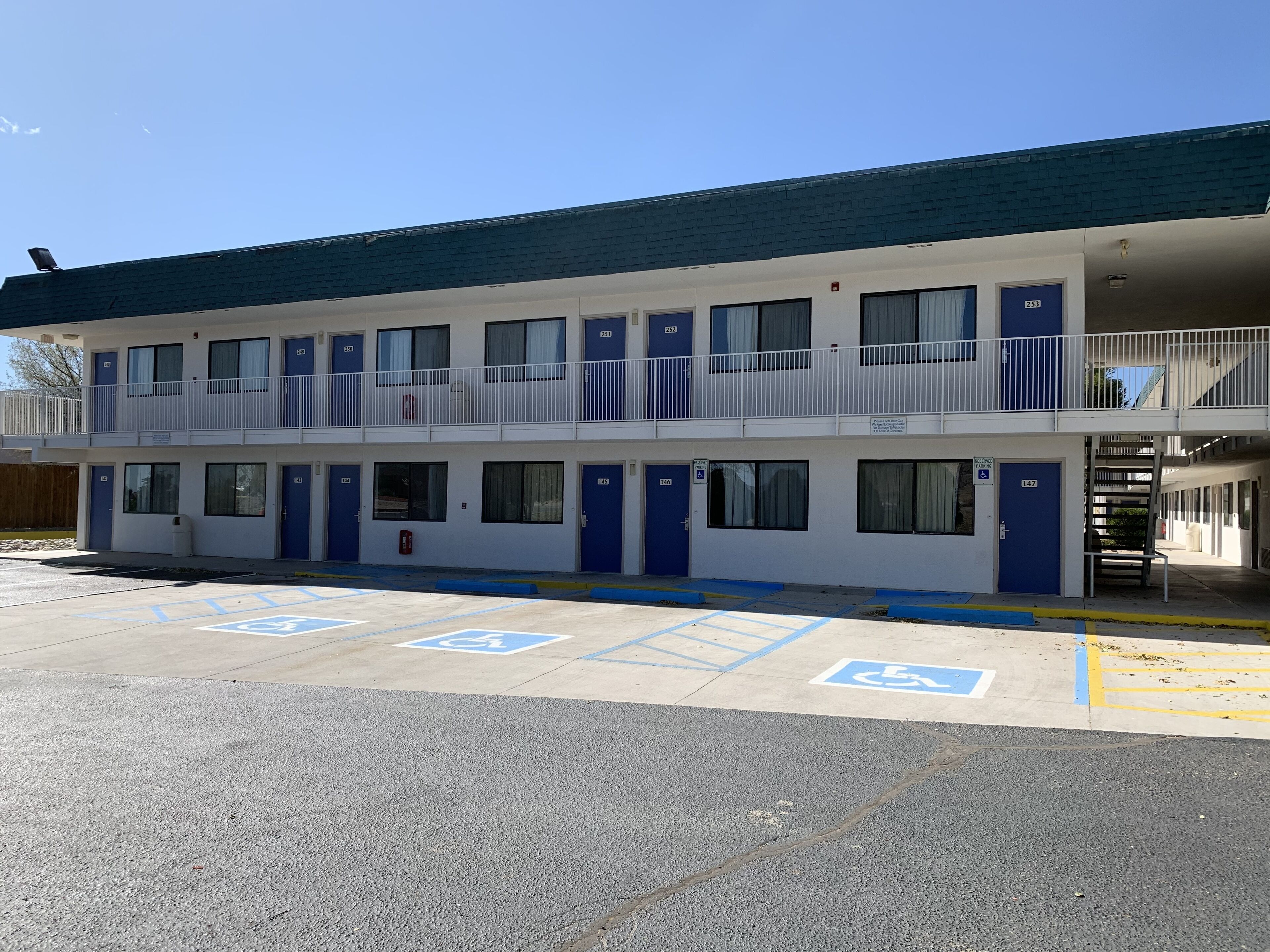 Un motel de dos pisos con puertas y ventanas azules, un área de estacionamiento techada y espacios de estacionamiento para discapacitados.