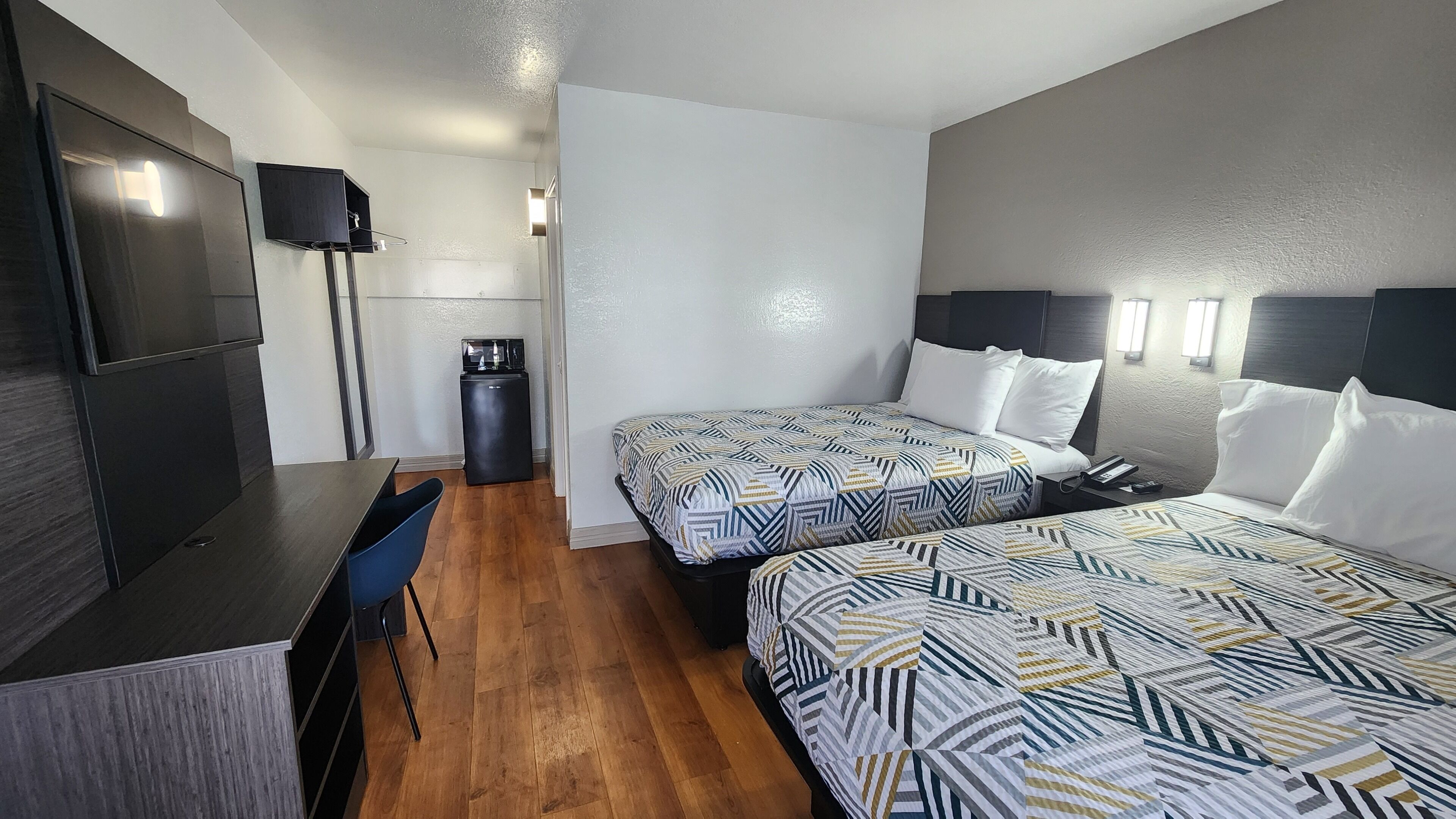 A hotel room with two beds, a desk, a chair, a small kitchenette, and a TV.