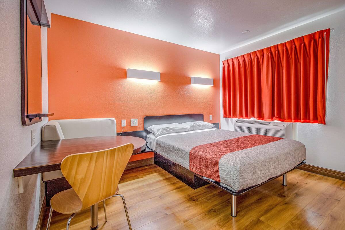 A hotel room with a bed, a desk, a chair, and a window with red curtains.