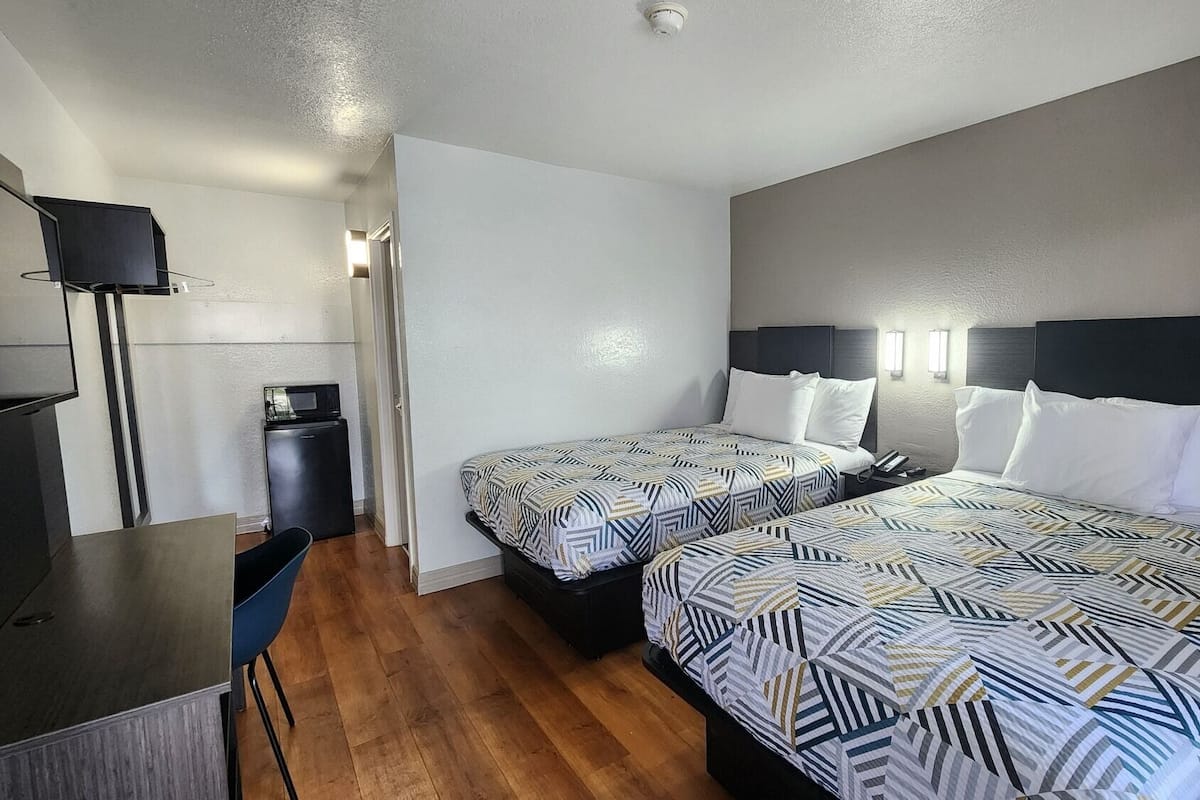 A hotel room with two beds, a desk, a television, and a small kitchenette.
