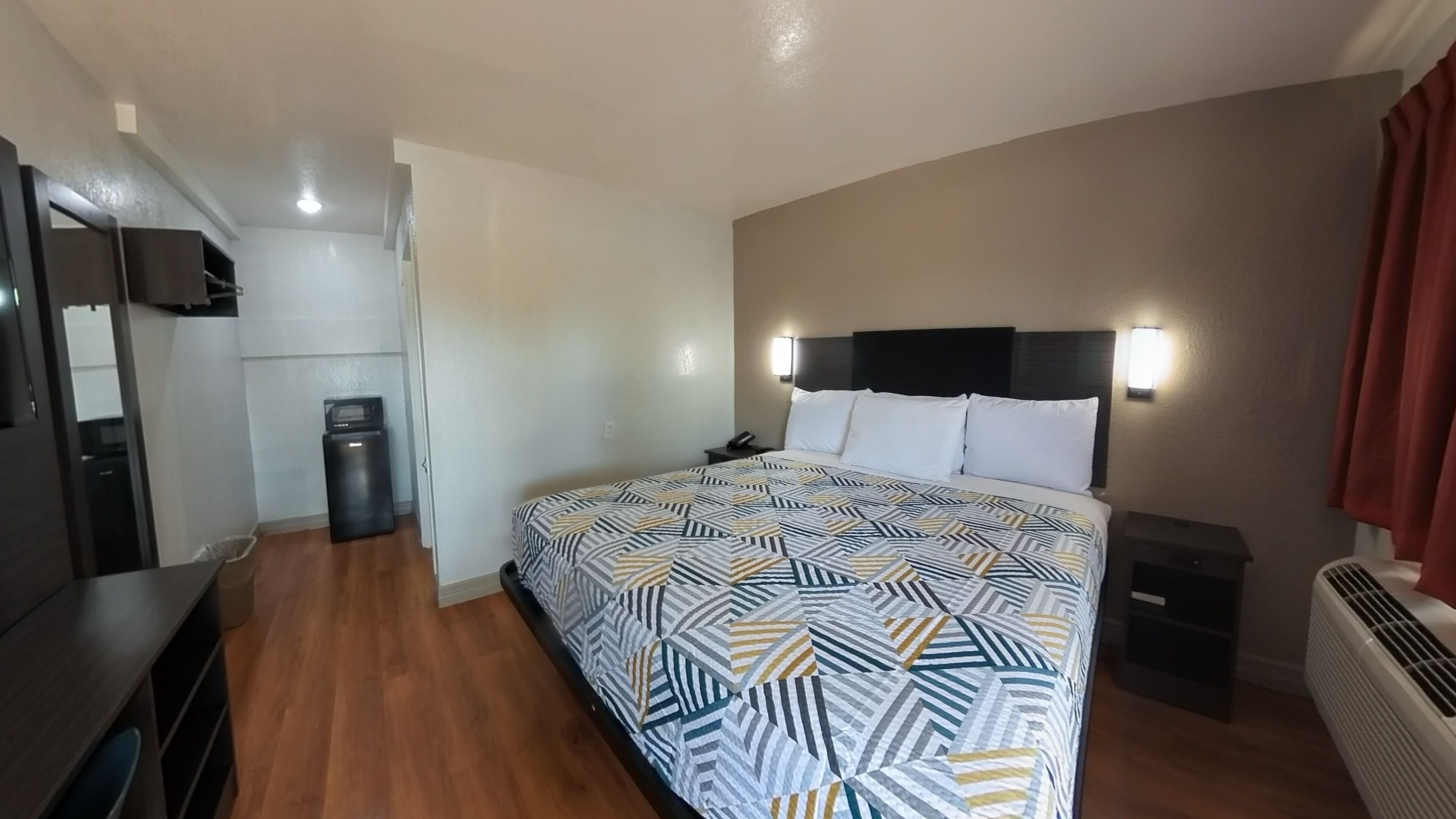 A hotel room with a bed, a desk, a chair, a heater, and a trash bin.