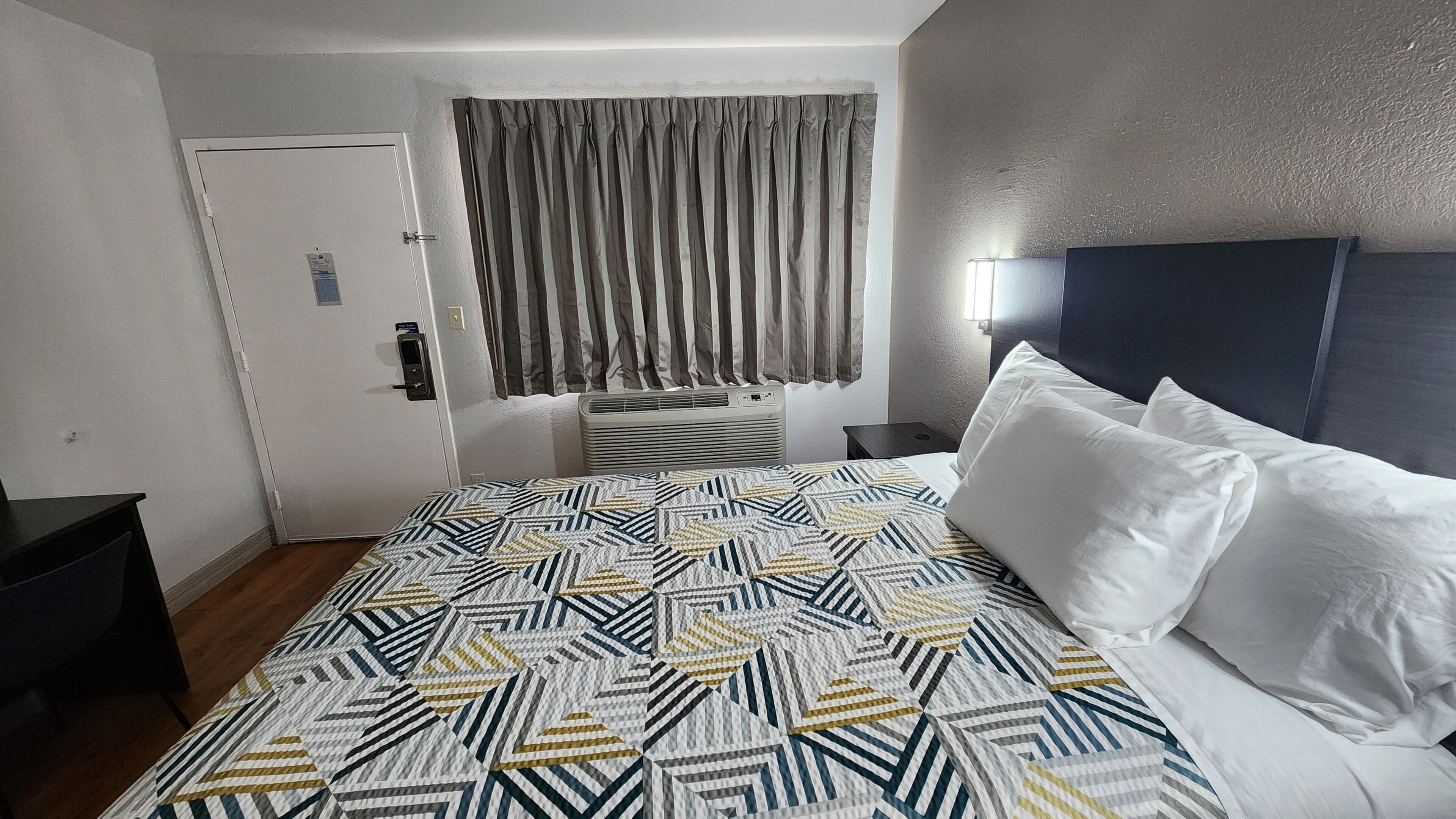 Ein Hotelzimmer mit Bett, Nachttisch, Tür und einem Fenster mit Vorhängen.