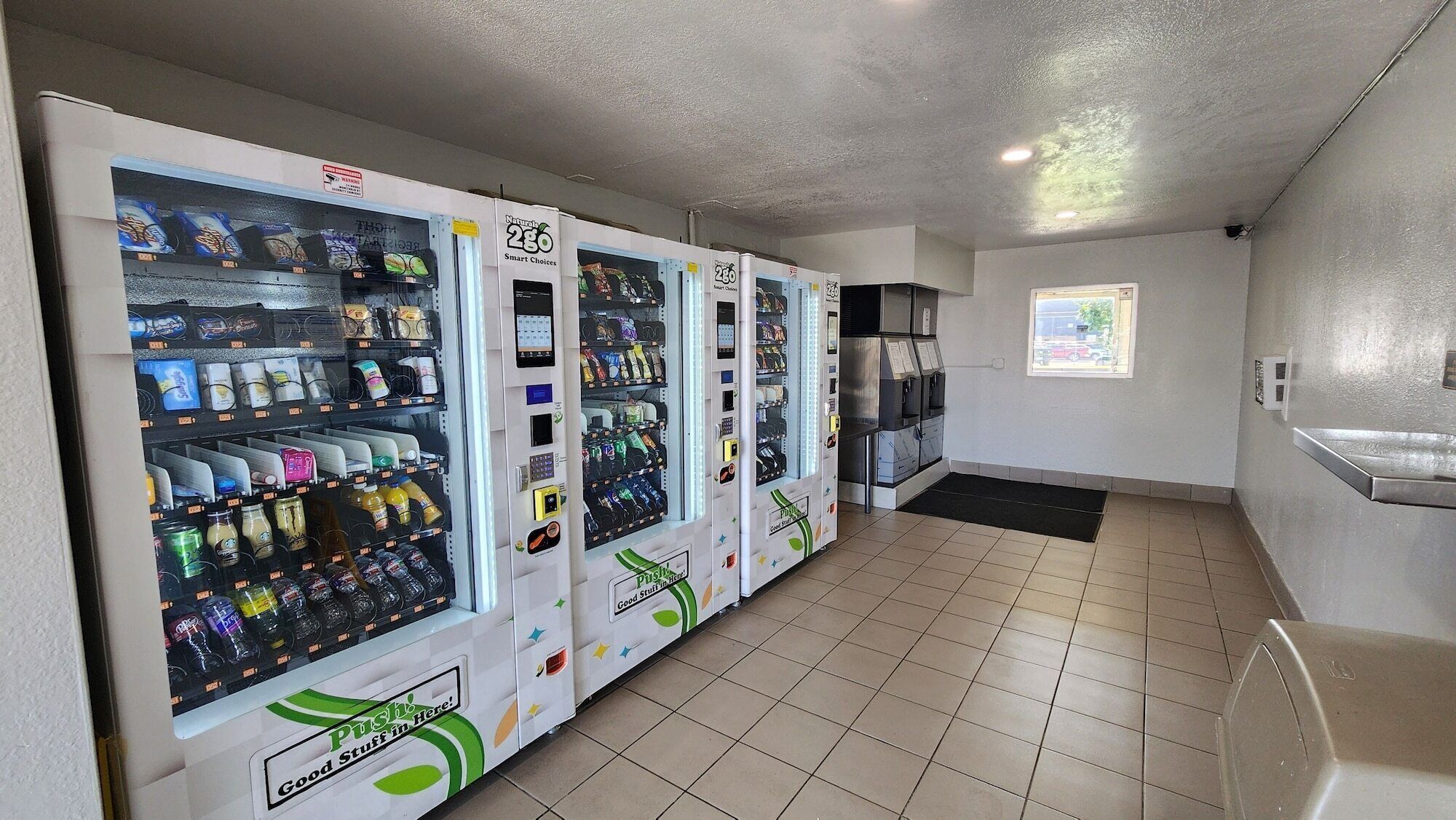 A row of vending machines with a variety of snacks and drinks, a tiled floor, and a window with a view of the outside.