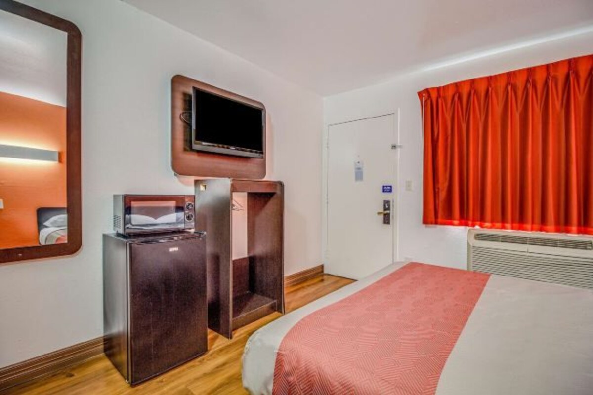 A hotel room with a bed, a TV mounted on a wooden panel, a minibar, and a red curtain.
