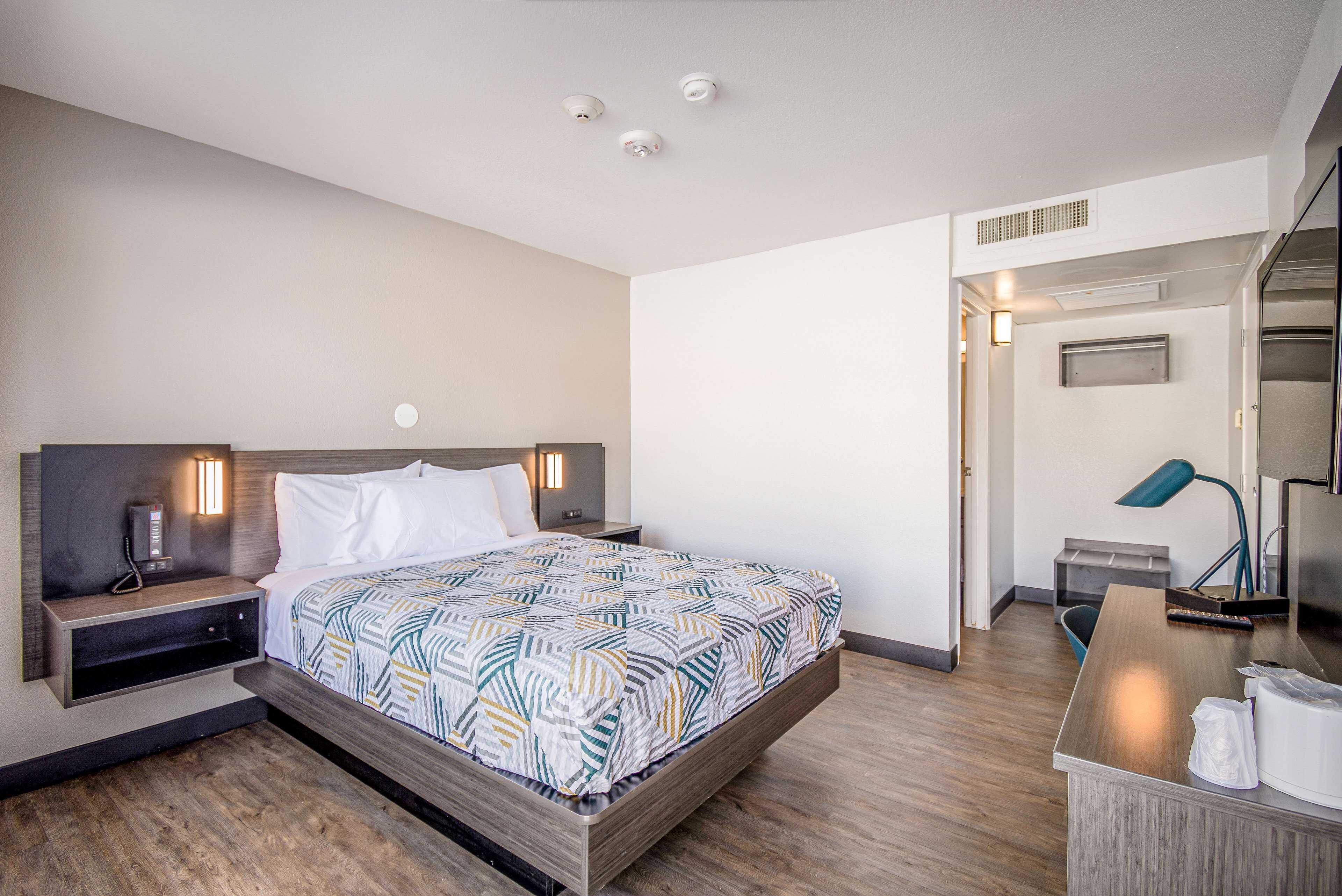 A modern hotel room with a large bed, bedside tables with lamps, a desk, and a kitchenette.