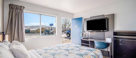 A hotel room with a large bed, a desk with a chair, a television, and a view of the outside.