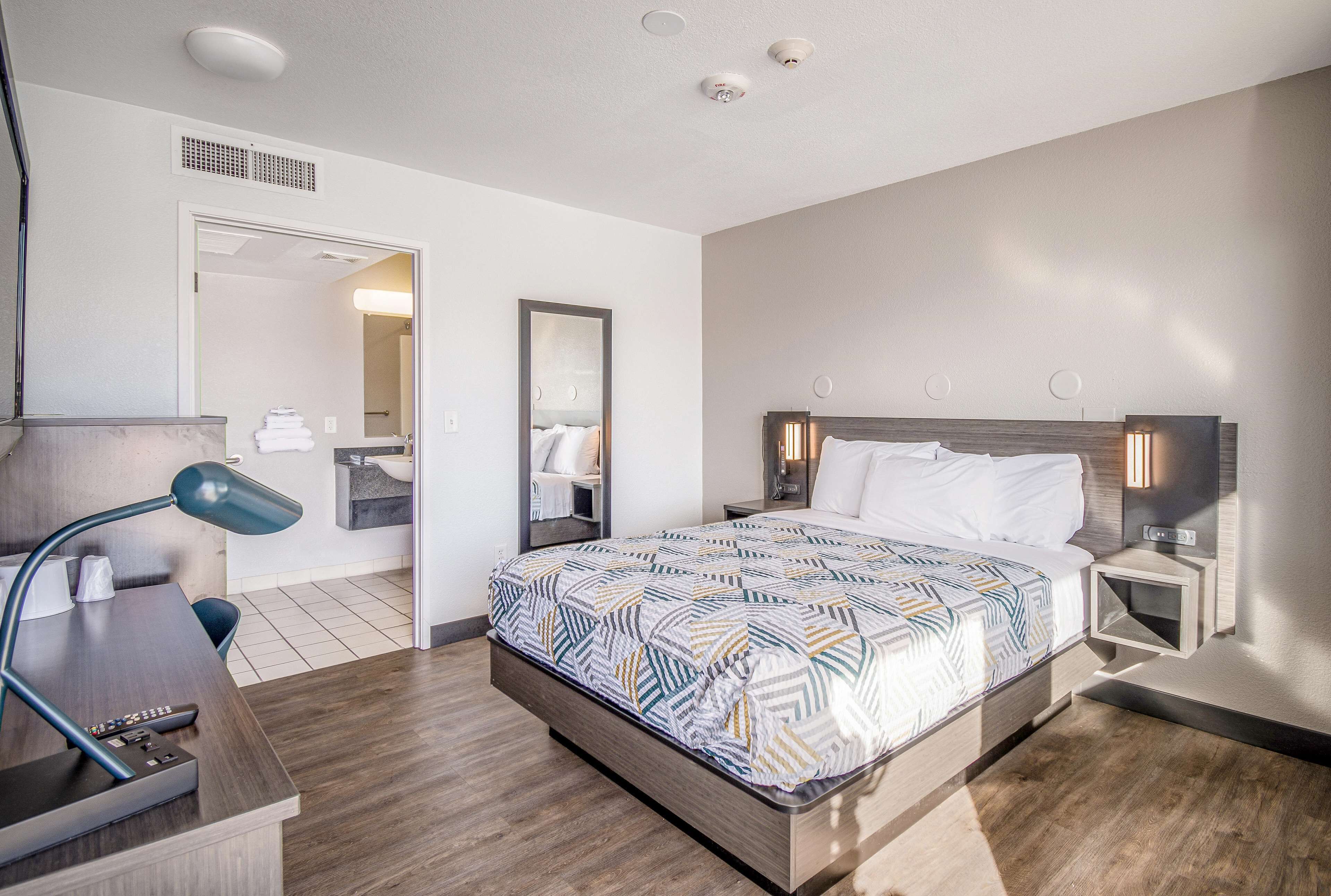 A hotel room with a bed, bedside tables, a desk with a lamp, a mirror, and a bathroom visible through an open door.