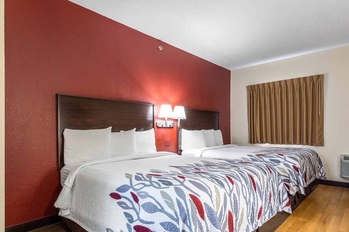 Hotellihuone, jossa on kaksi sänkyä, punainen seinä, puinen sängynpääty, kuviollinen päiväpeite ja ikkuna, jossa on verhot.