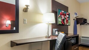 A hotel room with a desk, chair, lamp, television, coffee maker, and a coat rack. - Red Roof Inn Georgetown, IN - Louisville West (Georgetown)