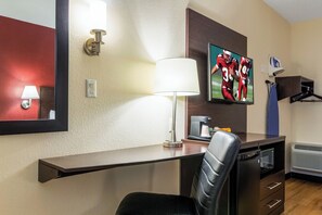 A hotel room with a desk, chair, lamp, television, coffee maker, and a coat rack. - Red Roof Inn Georgetown, IN - Louisville West (Georgetown)
