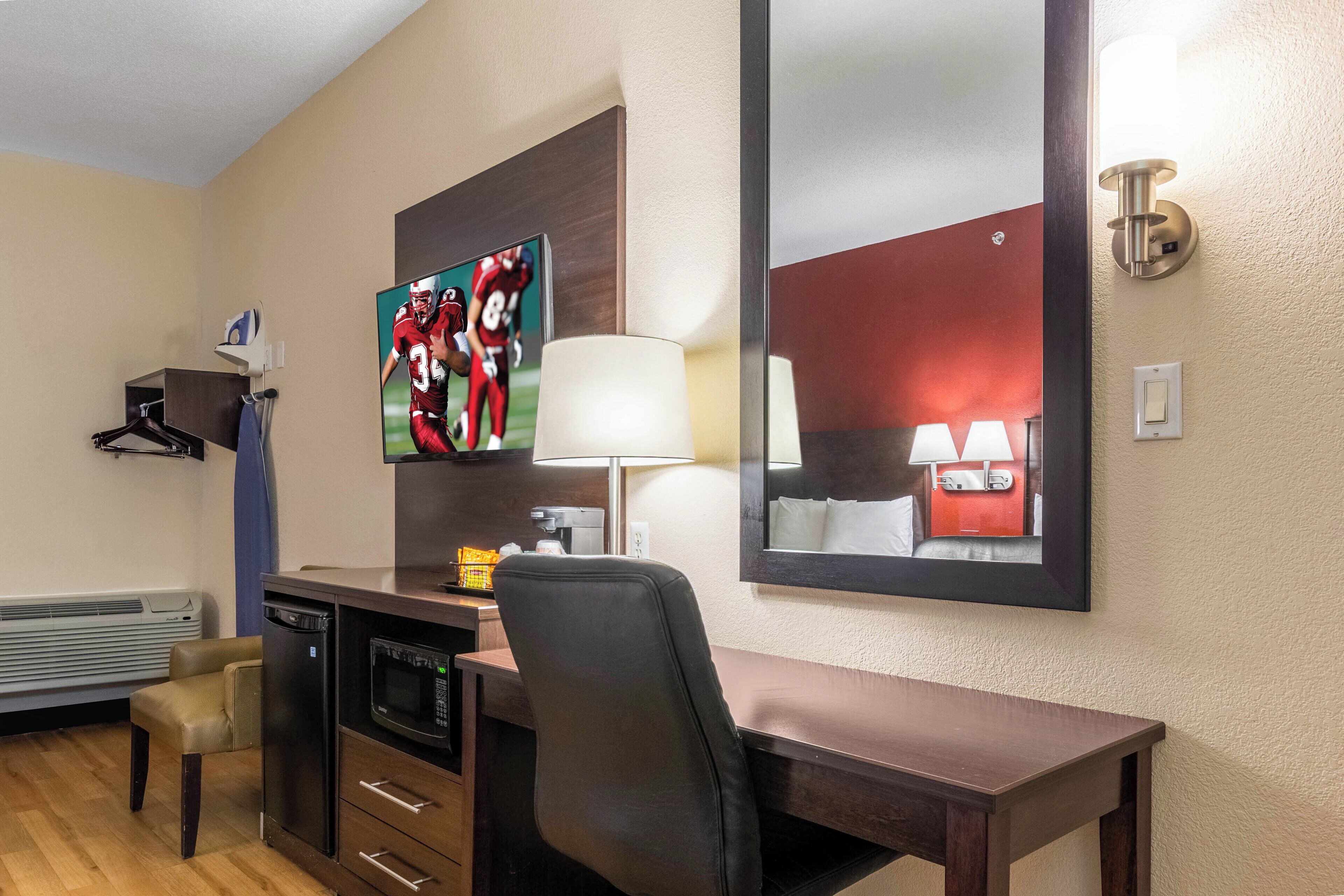 Habitación de hotel con escritorio, silla, televisor, lámpara y un espejo grande.