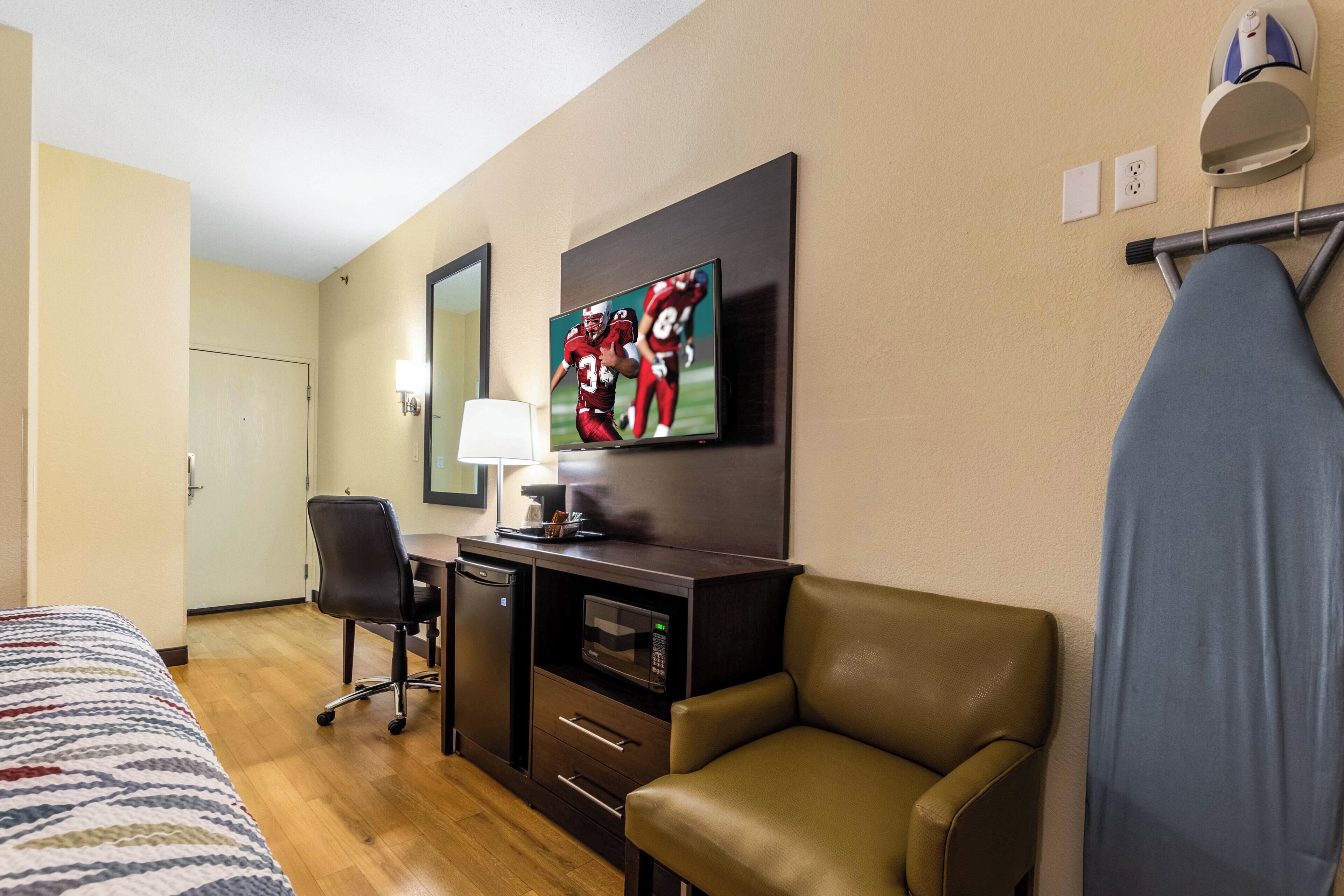 Una habitación de hotel con una cama, un escritorio con computadora, una silla, un televisor montado en la pared y una ventana con cortinas.