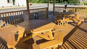 A wooden deck with picnic tables and benches, a trash can, and a building in the background. - Red Roof Inn Georgetown, IN - Louisville West (Georgetown)