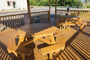 Una terraza de madera con mesas de picnic, bancos, un bote de basura y un edificio al fondo.
