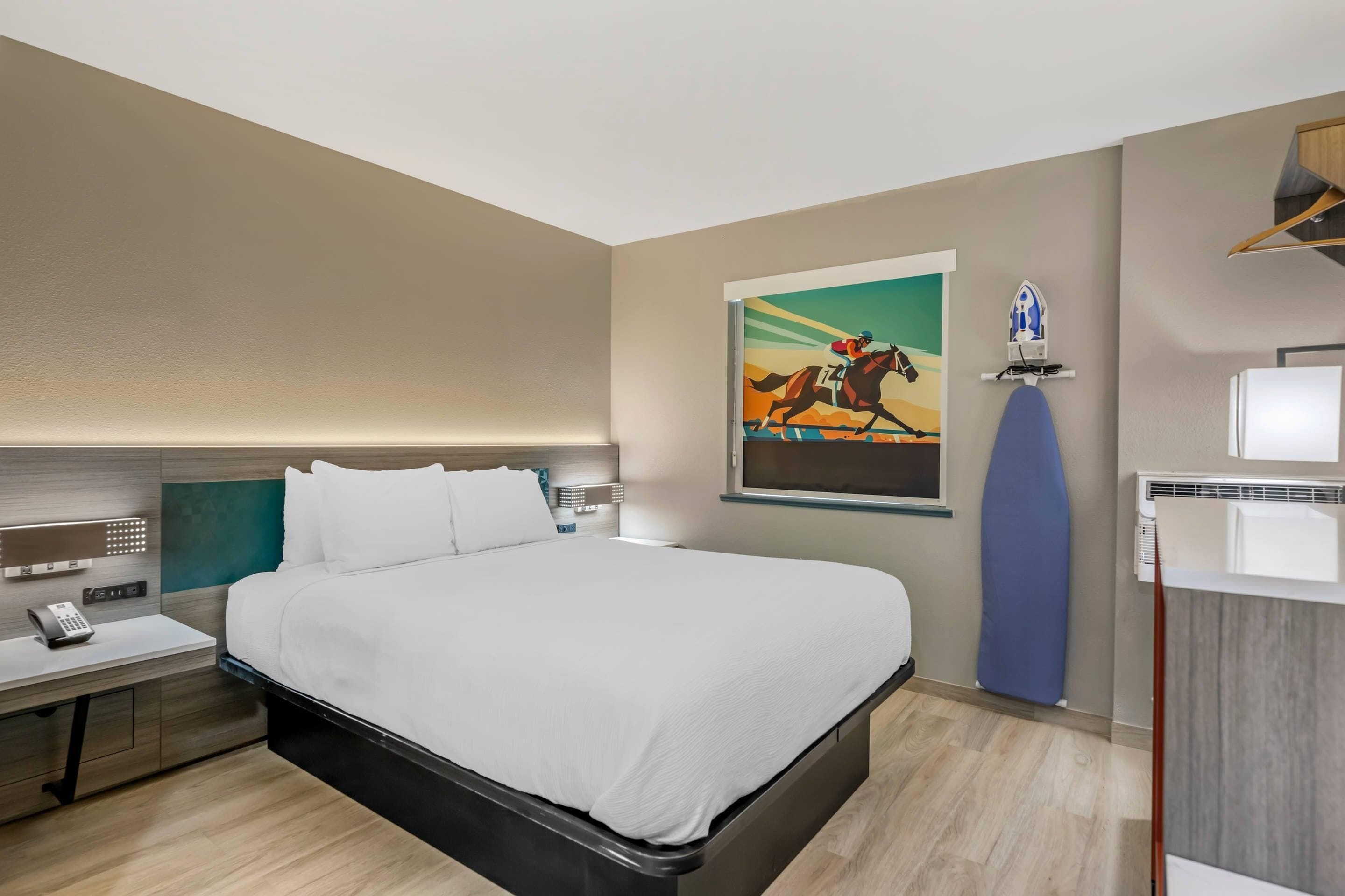 Habitación de hotel con una cama grande, una mesita de noche con un teléfono, una tabla de planchar en la pared y un baño pequeño con espejo y lavabo.