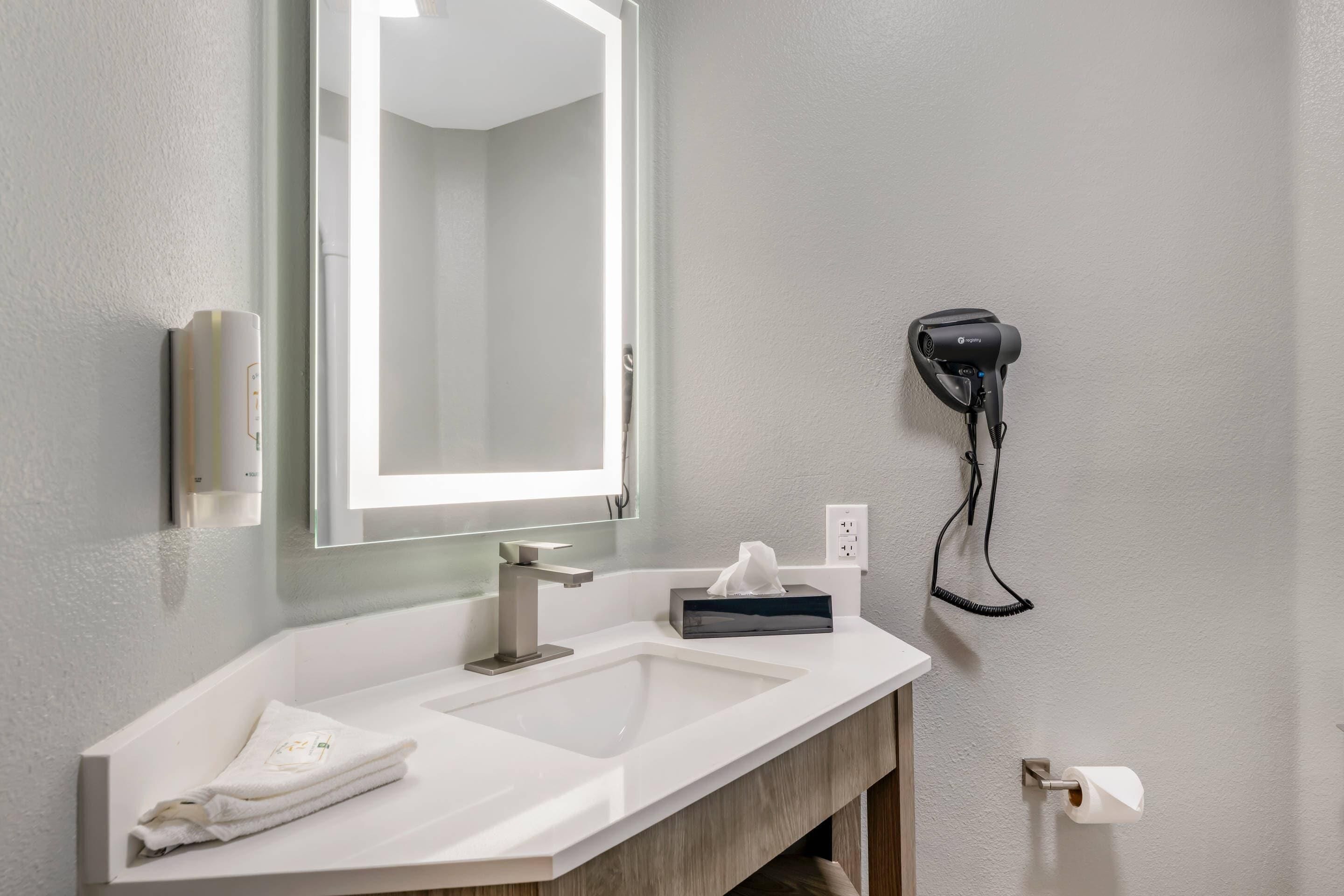 Une salle de bain avec un lavabo blanc, un miroir, un sèche-cheveux fixé au mur et une serviette posée sur le comptoir.