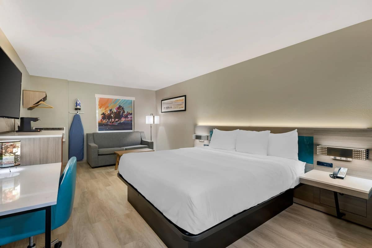 A modern hotel room with a large bed, a desk with a chair, a small sofa, and a bedside table with a phone.