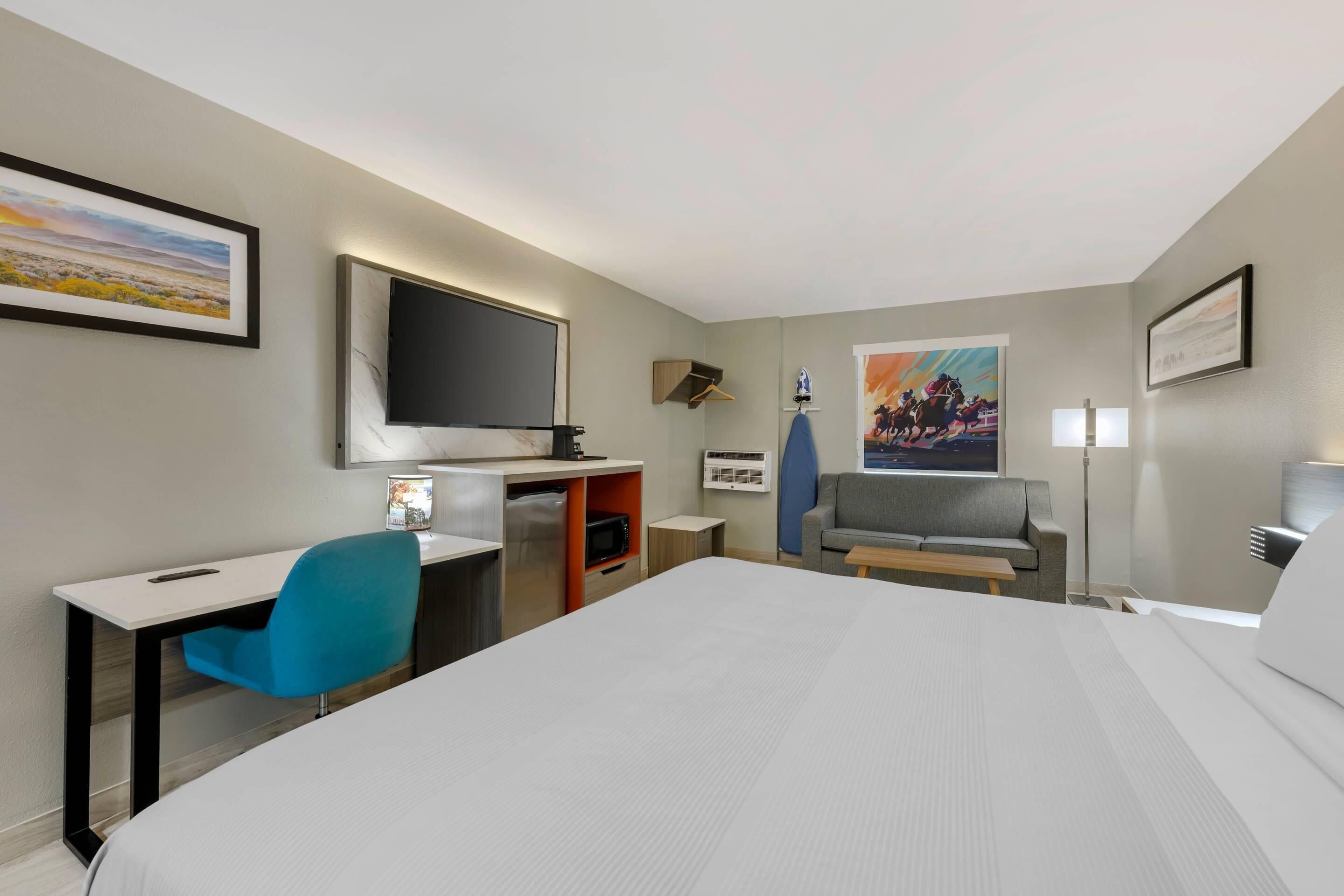 A hotel room with a bed, a desk with a chair, a television, a sofa, and artwork on the walls.
