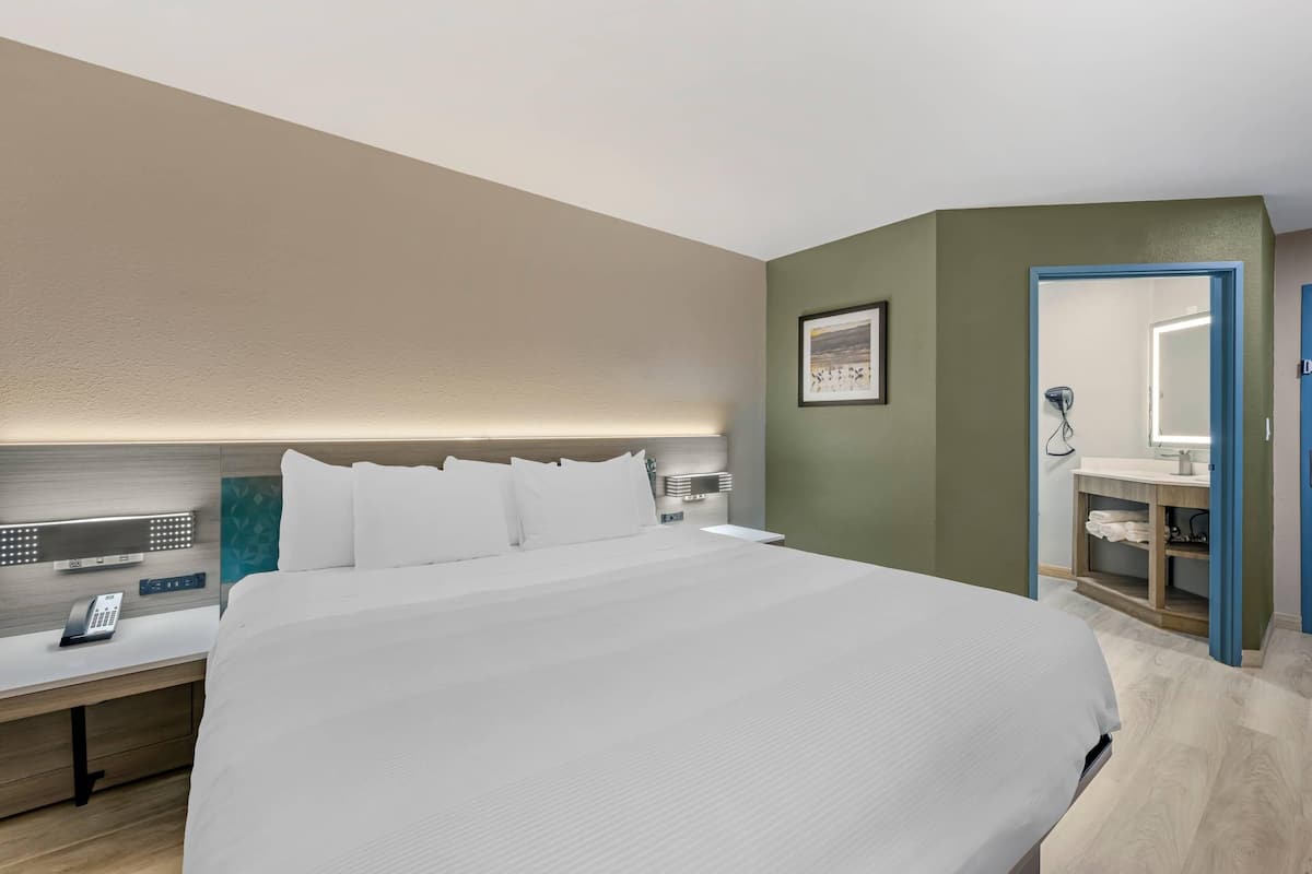 A hotel room with a large bed, bedside table with a phone, a wall-mounted light, and a bathroom with a sink and mirror.
