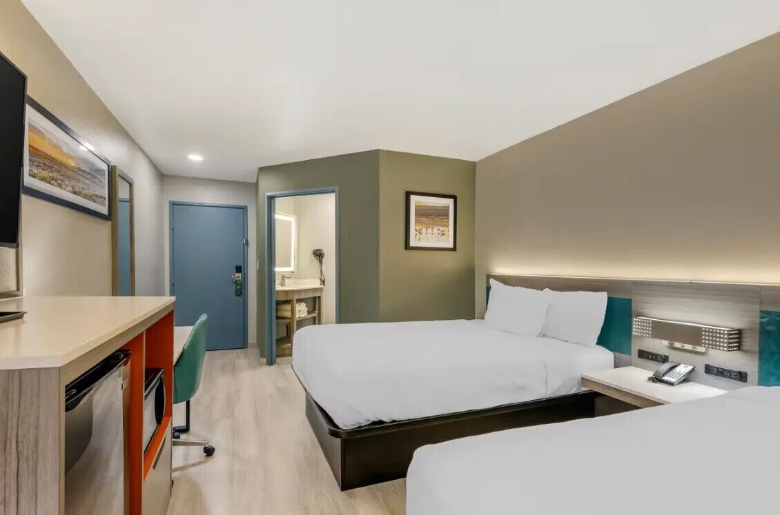 A hotel room with two beds, a desk with a chair, a wardrobe, and a bathroom visible through an open door.