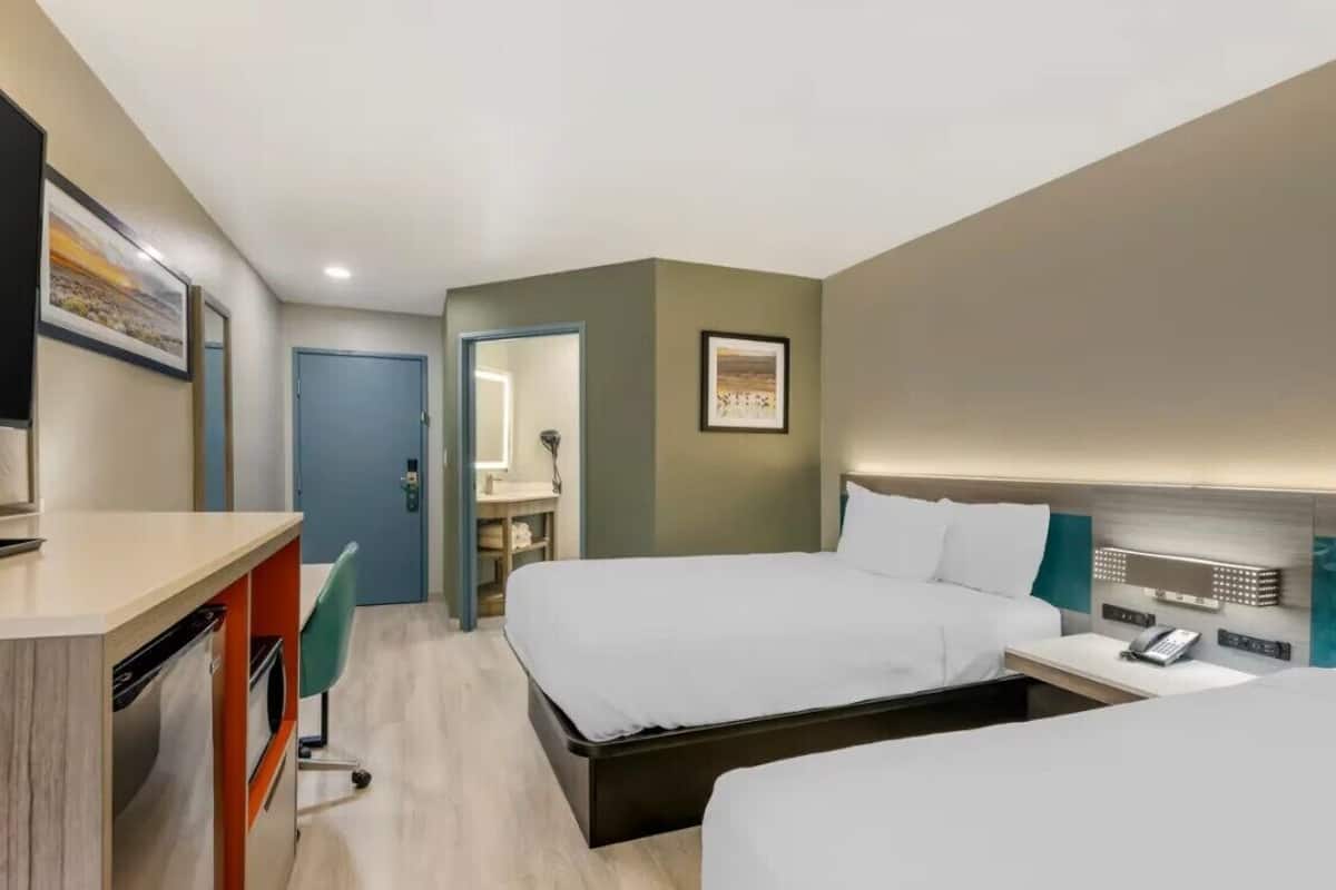 A hotel room with two beds, a desk with a chair, a wardrobe, and a bathroom visible through an open door.
