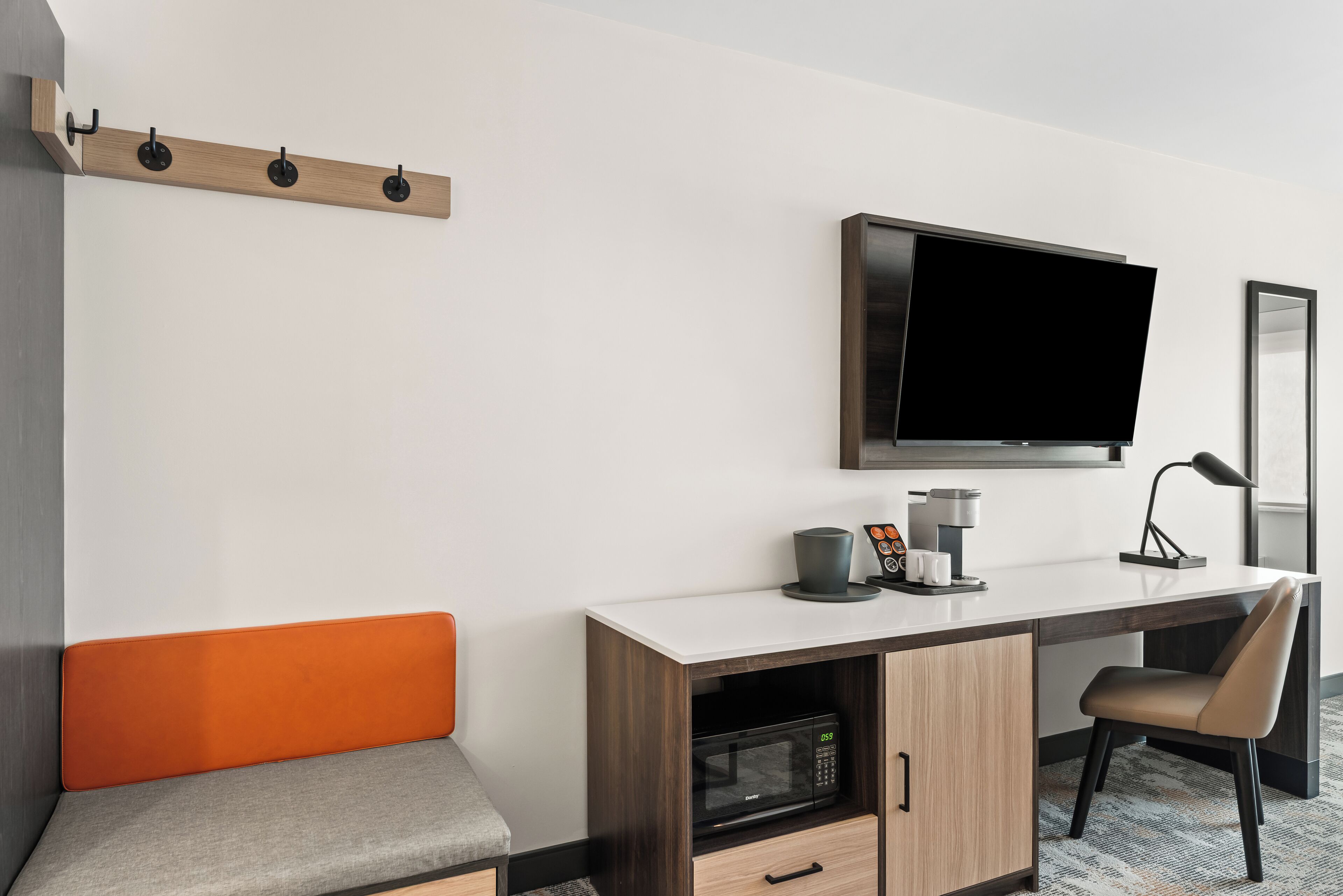 Habitación de hotel moderna con televisión de pantalla plana, un escritorio con cafetera, microondas y un banco acolchado.
