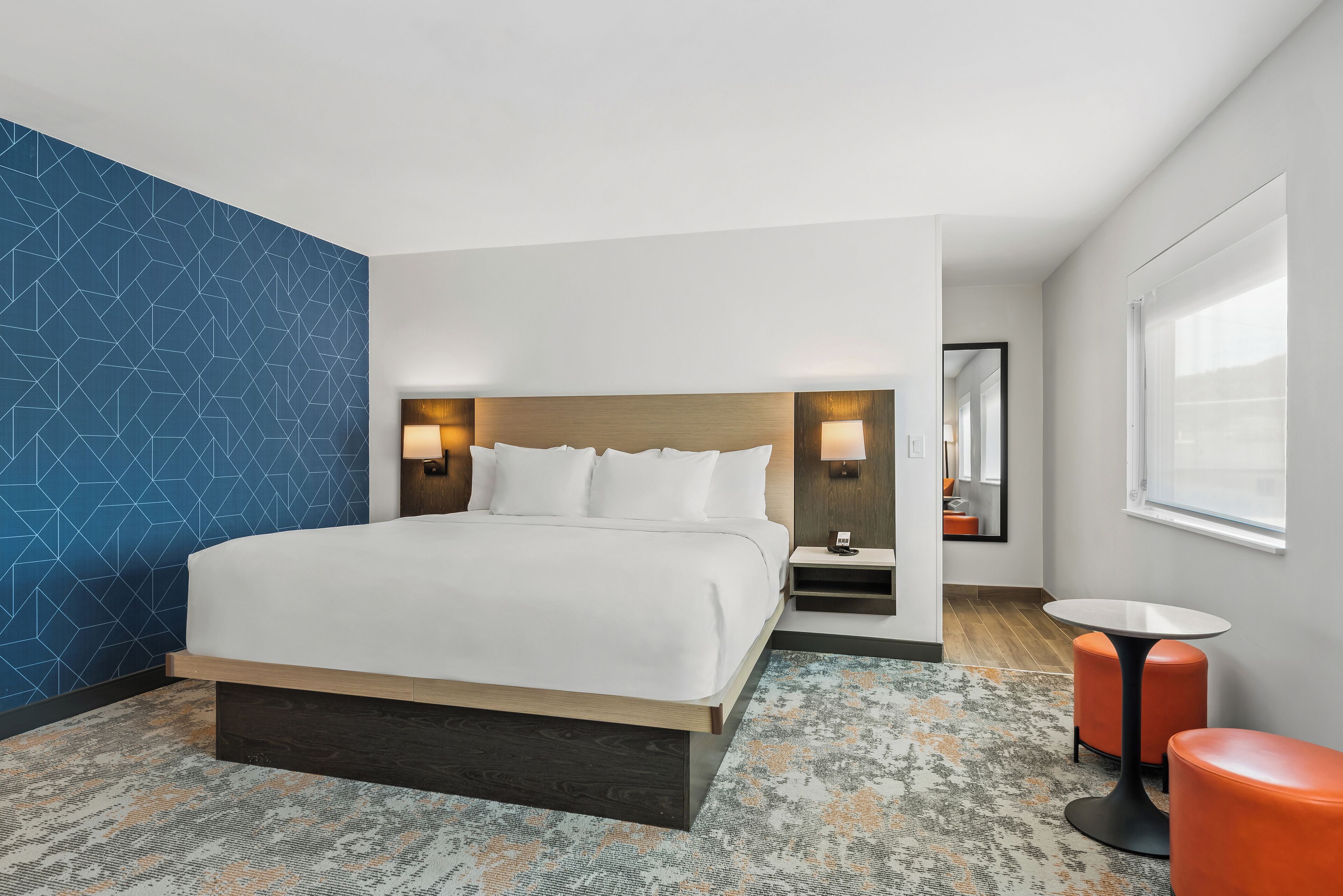 A modern hotel room with a large bed, a small table, two orange stools, and a large window.