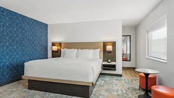 A modern hotel room with a large bed, a small table, two orange stools, and a large window.