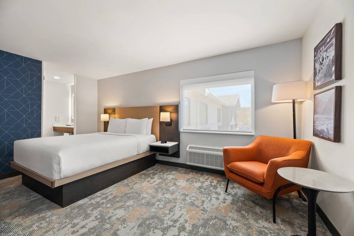 Ein Hotelzimmer mit Bett, Sessel, Tisch, Nachttisch, Lampe, Fenster und einem Wandbild.