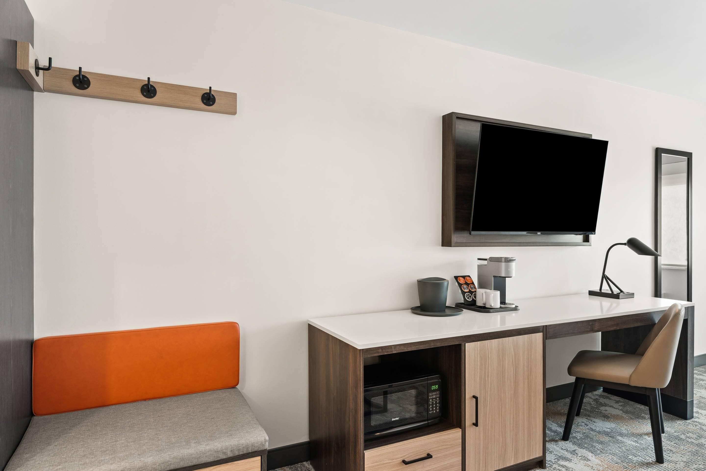 Una habitación de hotel moderna con televisión de pantalla plana, escritorio con cafetera, microondas y un banco acolchado.