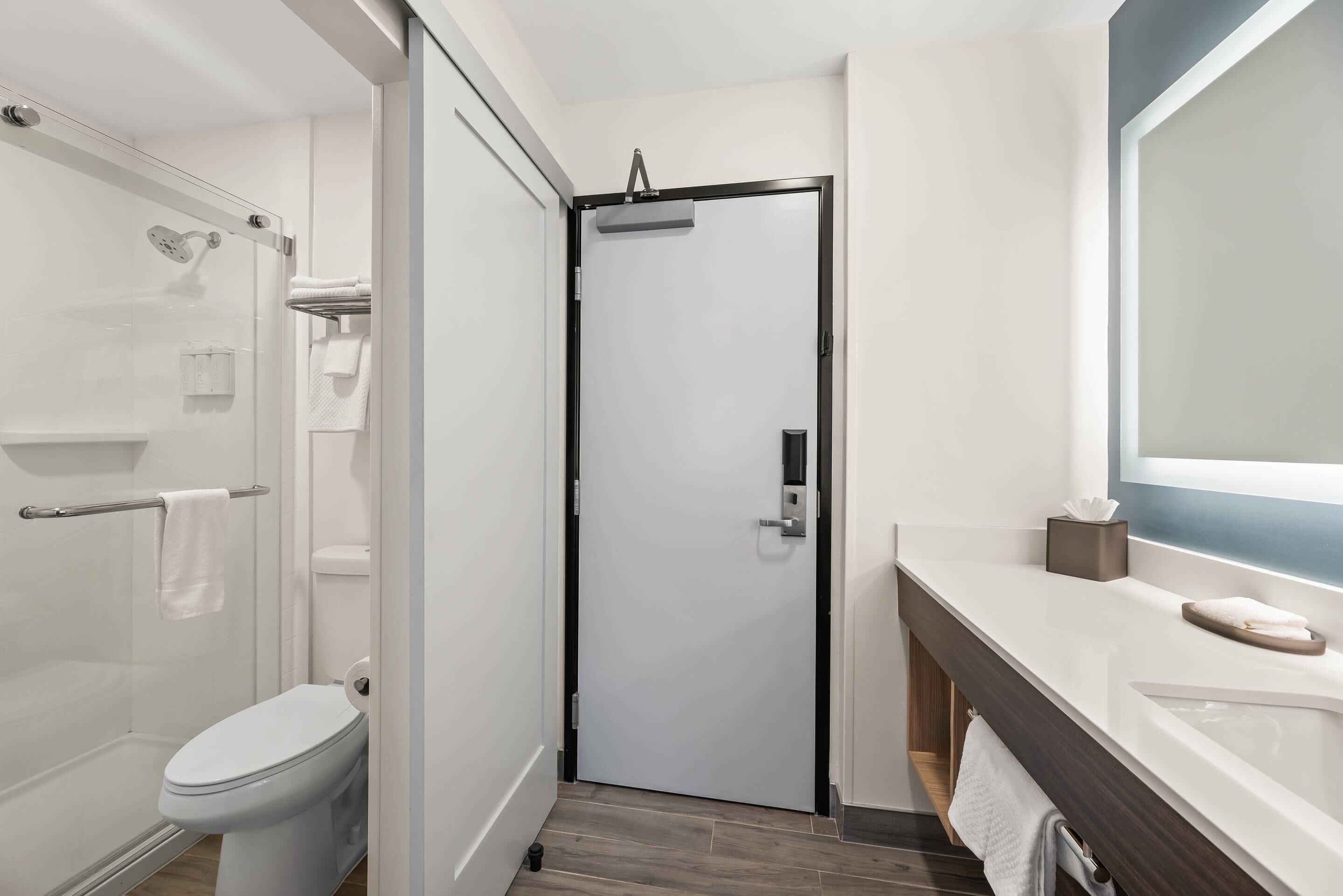 Un baño moderno con cabina de ducha de vidrio, inodoro blanco, tocador de madera oscura y un espejo grande.