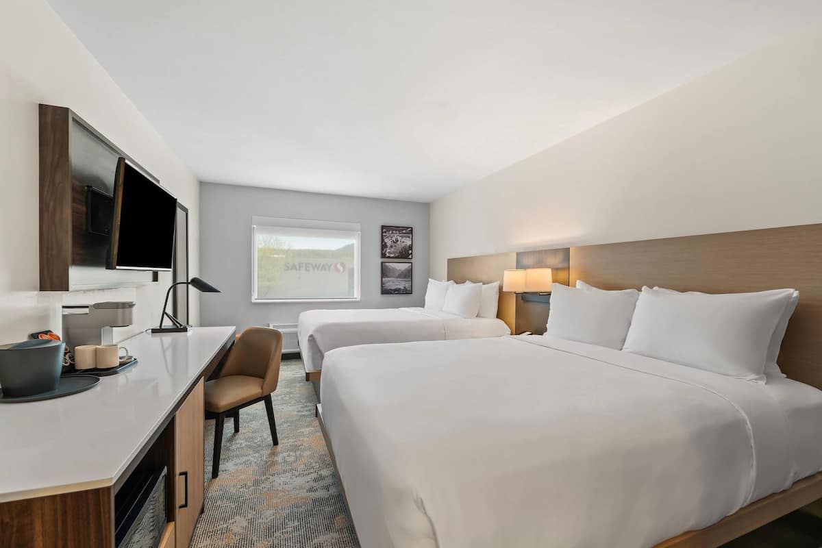 Ein Hotelzimmer mit zwei Betten, einem Schreibtisch, einem Flachbildfernseher, einem Sessel, einer Lampe und einem Fenster mit Landschaftsblick.