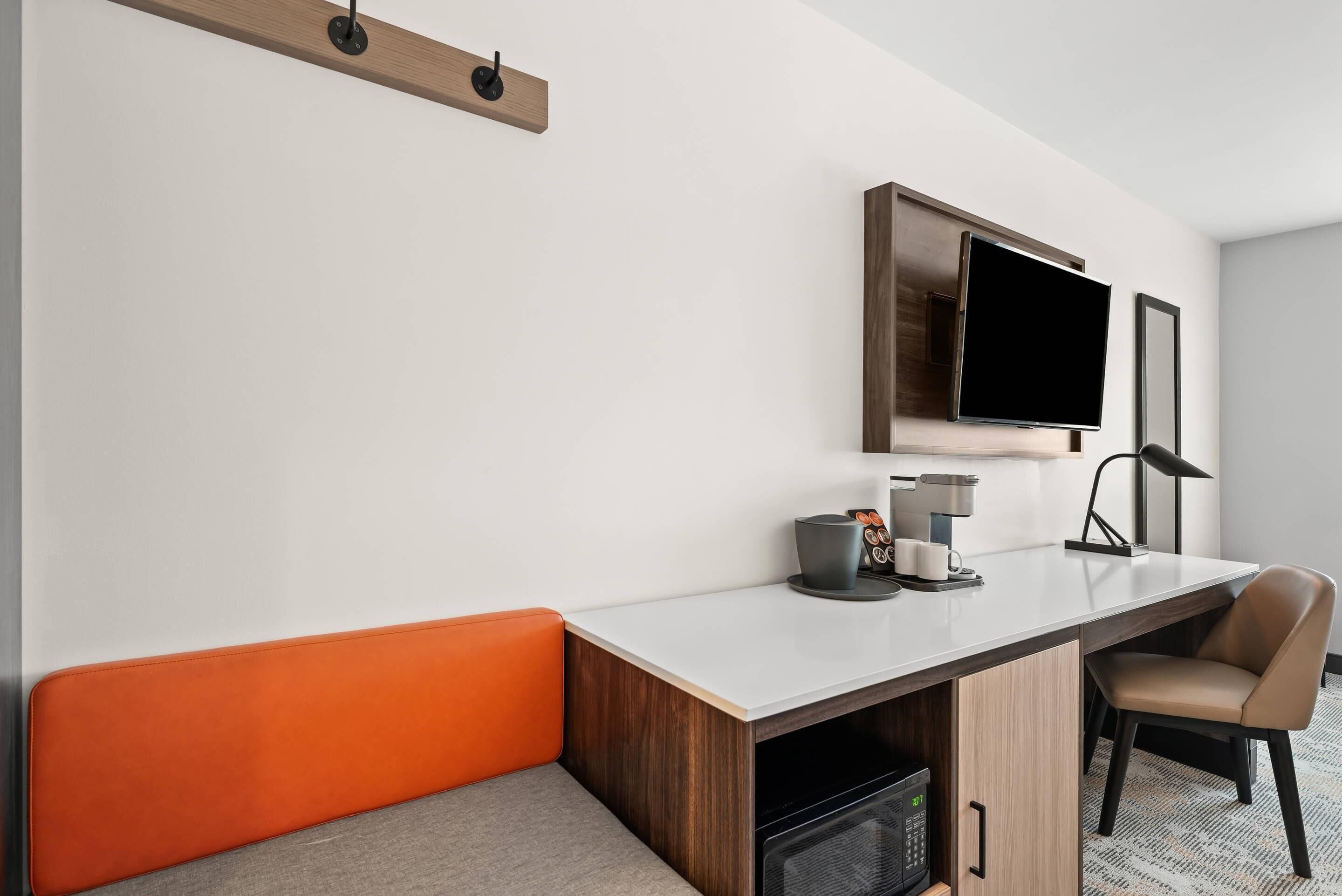 A modern hotel room with a white wall, a flat-screen TV mounted above a desk, a chair, a coffee maker, and a small cabinet.