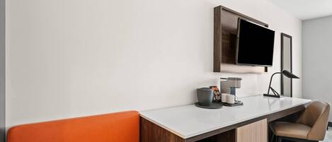 A modern hotel room with a white wall, a flat-screen TV mounted above a desk, a chair, a coffee maker, and a small cabinet.