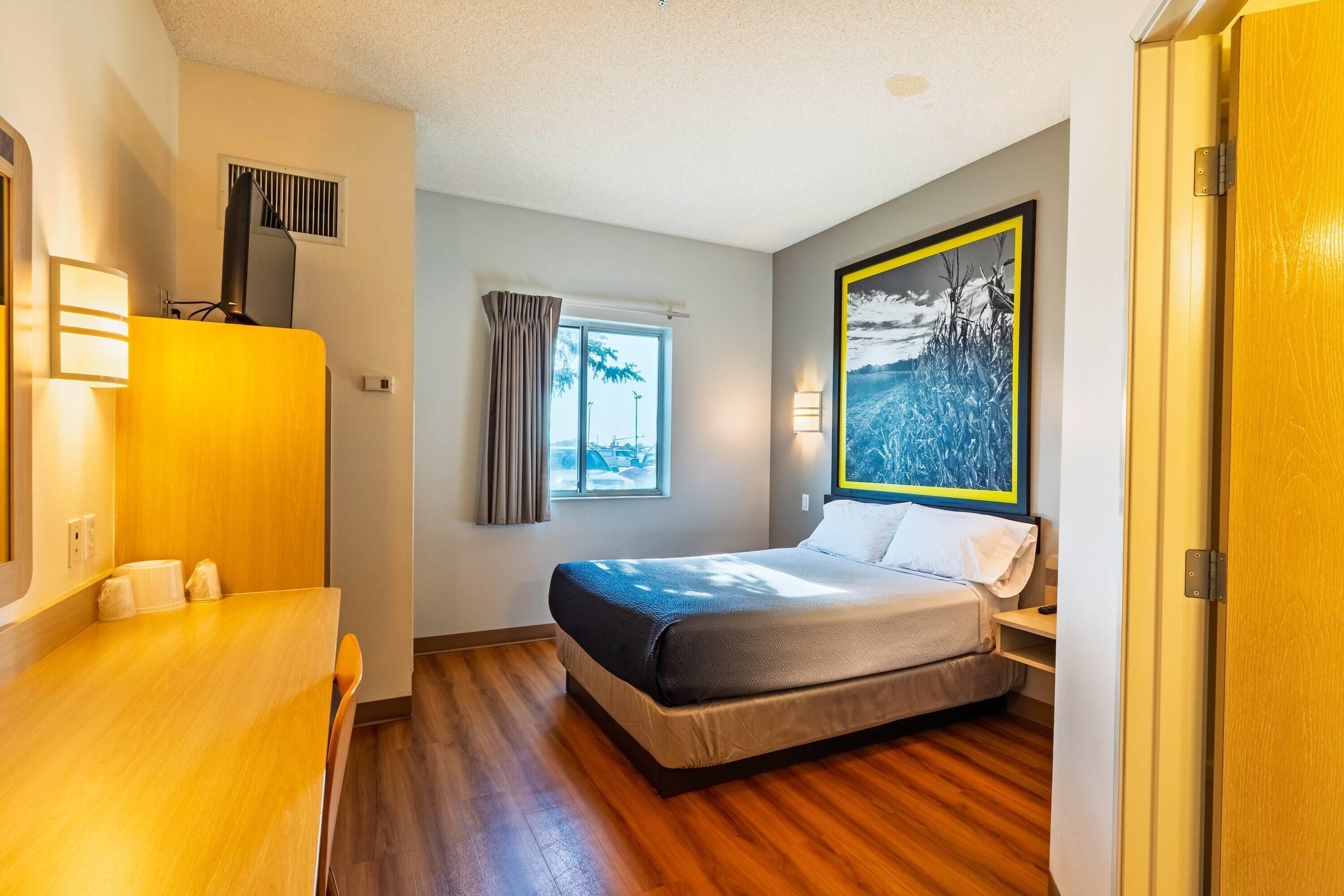 Quarto de hotel com cama, escrivaninha, janela com cortinas, um quadro e uma porta.