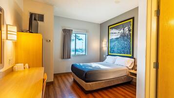 Quarto de hotel com cama, escrivaninha, janela com cortinas, um quadro e uma porta.