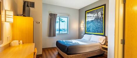 Una habitación de hotel con cama, escritorio, ventana con cortinas, un cuadro y una puerta.