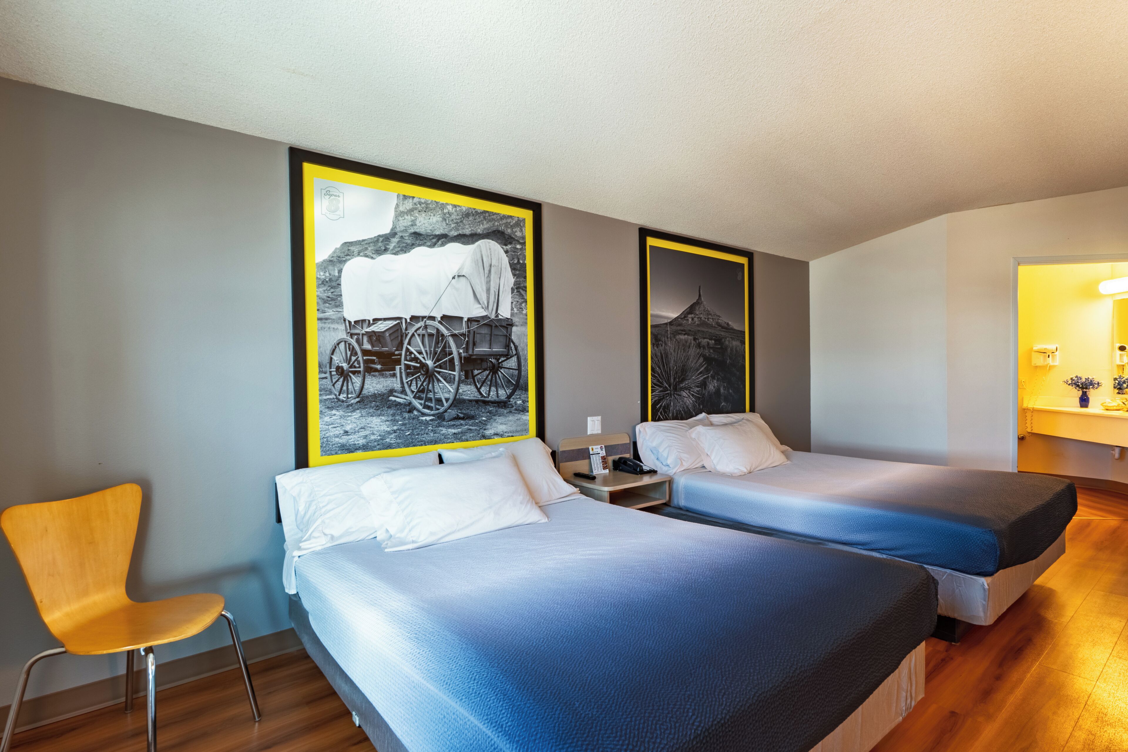 Habitación de hotel con dos camas, una silla, una mesita de noche y dos cuadros en la pared.