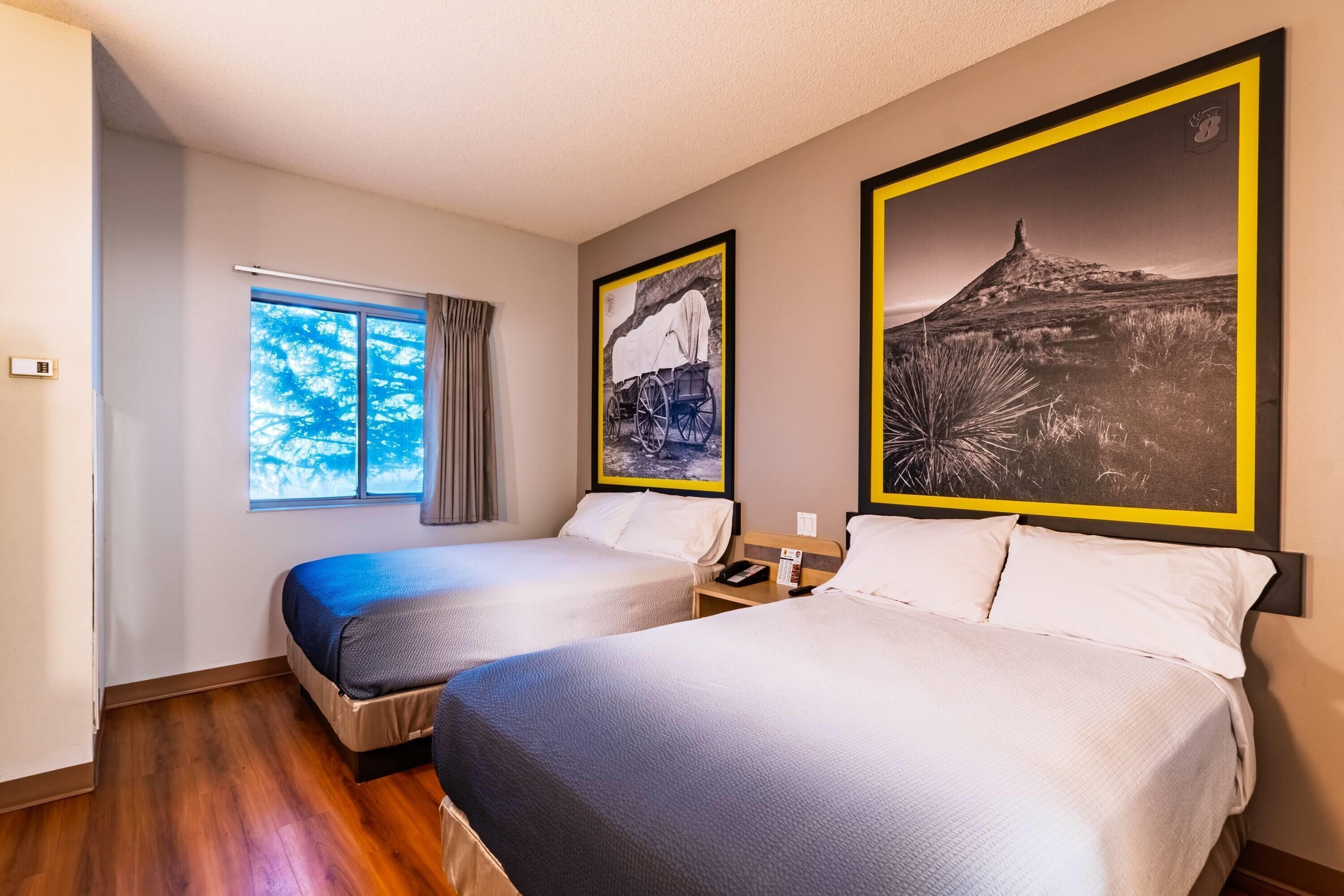 A hotel room with two beds, a window with curtains, and two framed pictures on the wall.