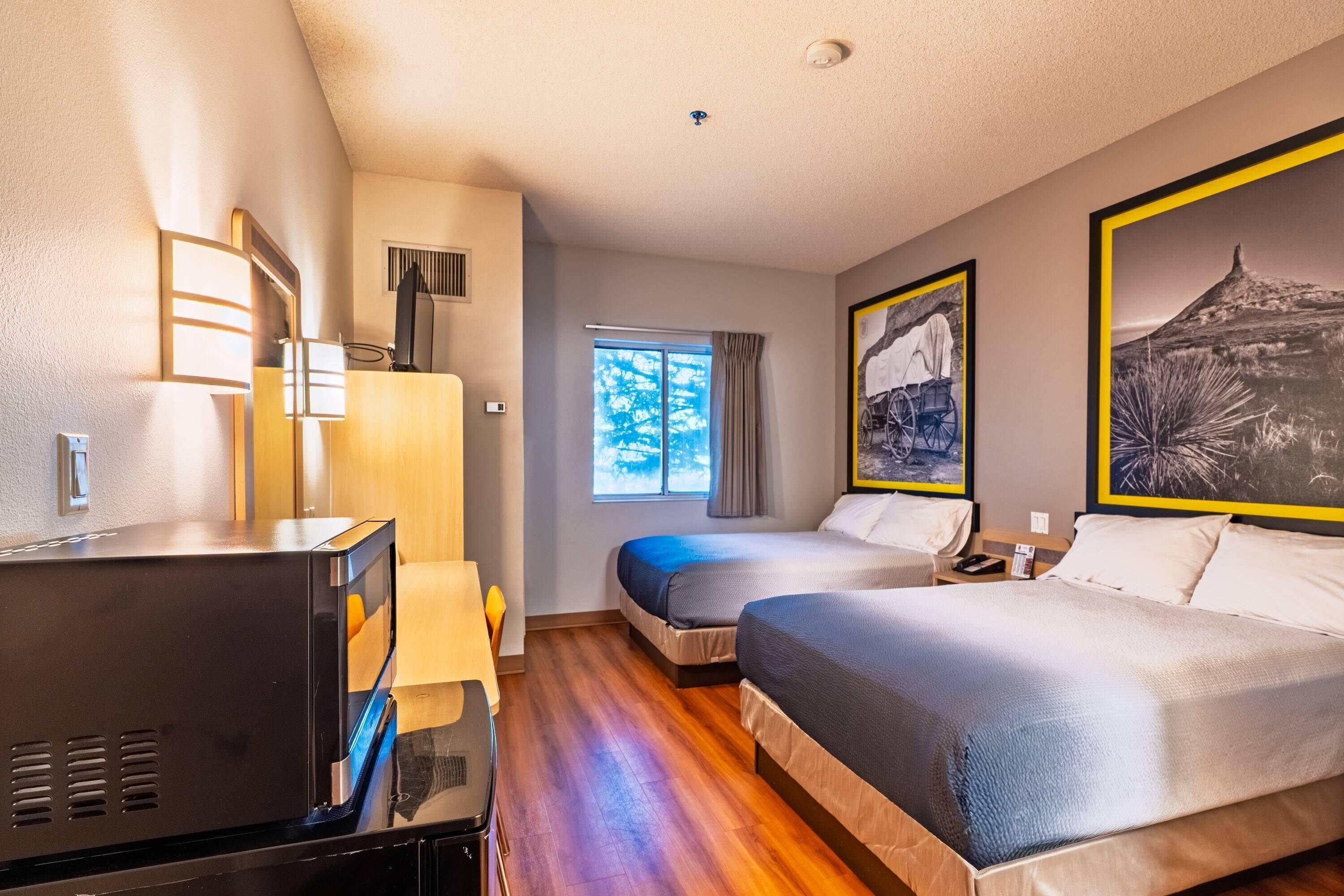 A hotel room with two beds, a desk with a television, a window with curtains, and artwork on the walls.