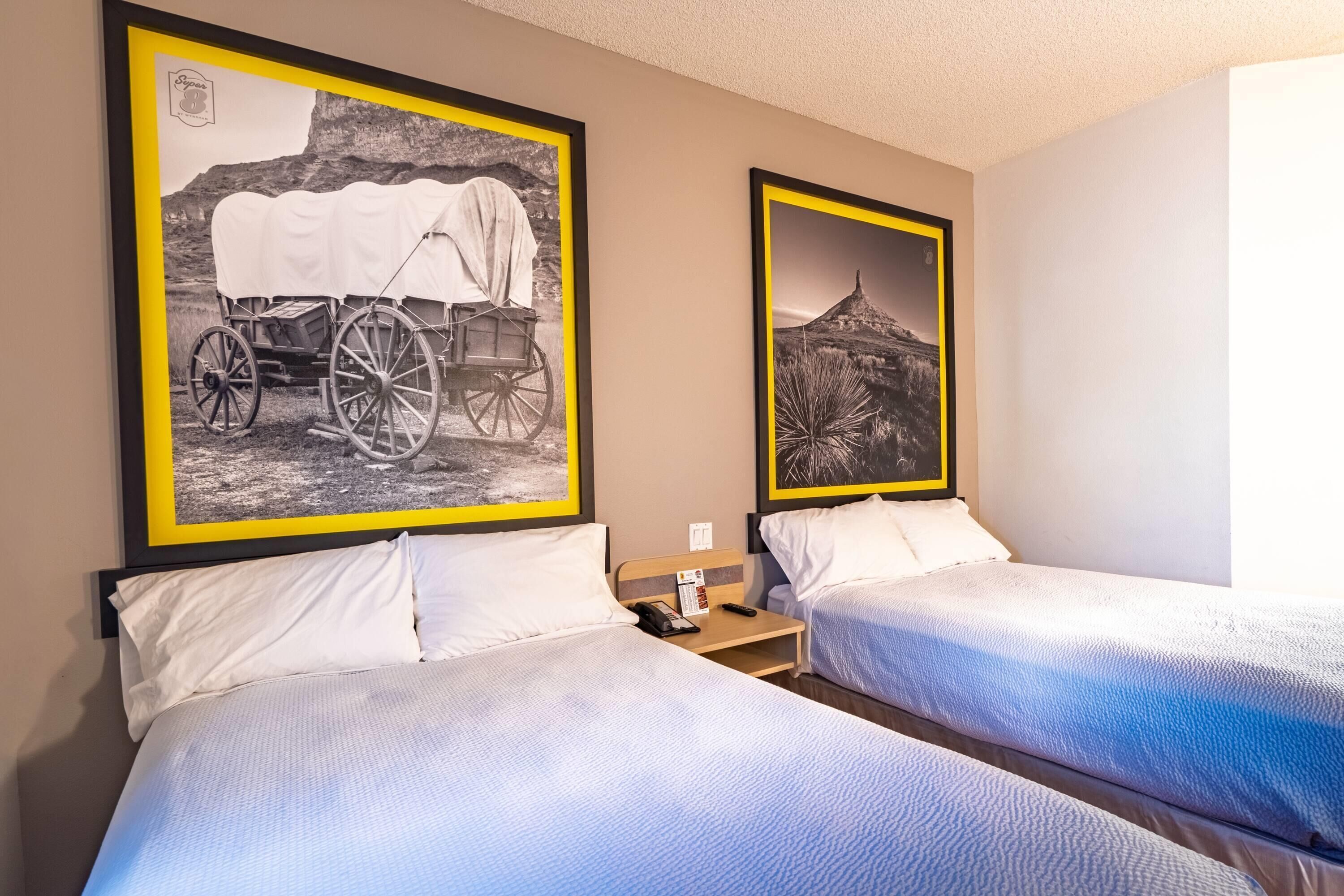 A hotel room with two beds, each with white bedding and a headboard. There are framed black and white photographs on the wall above the beds.